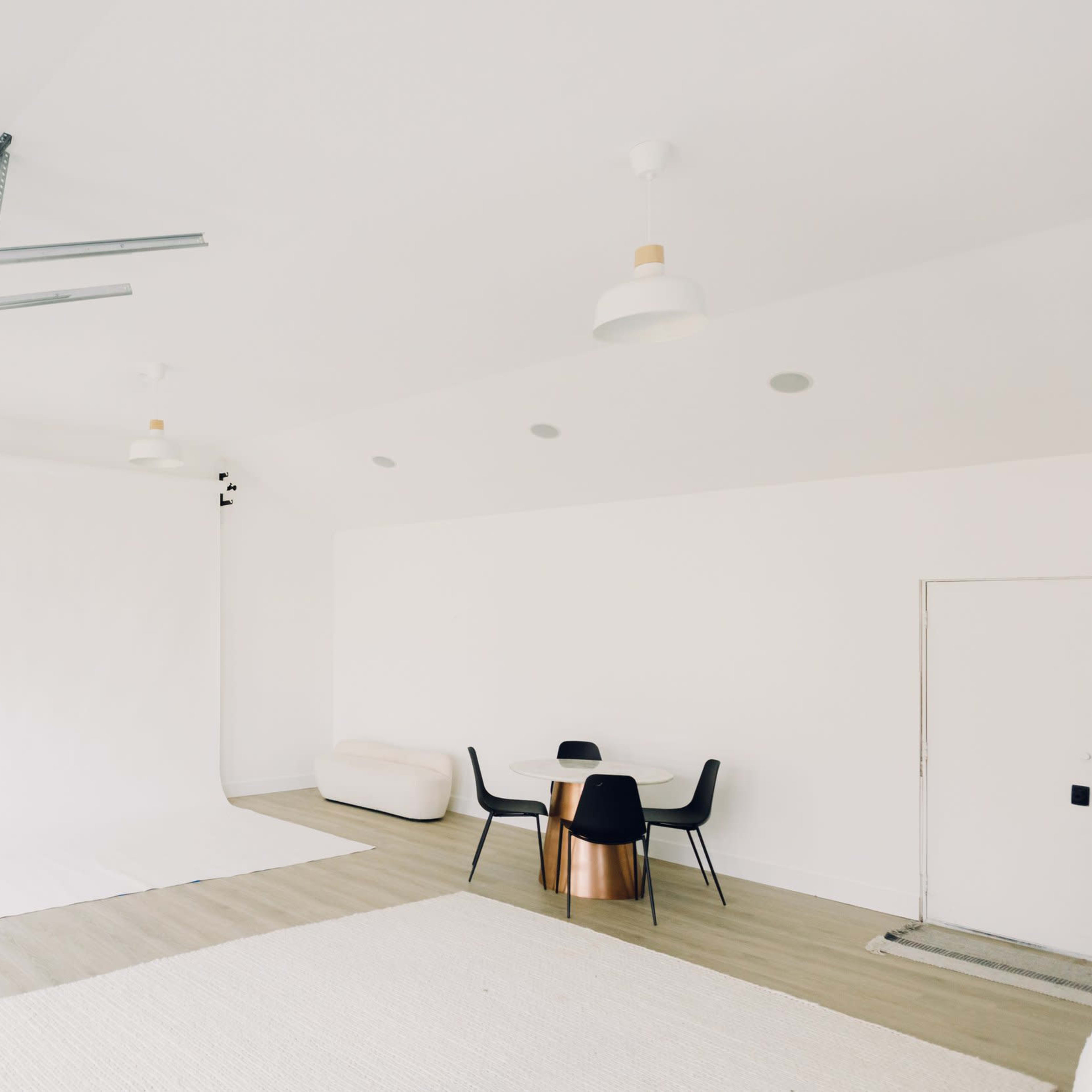 A minimalist studio space with a white backdrop, a round wooden table with black chairs, and a light-colored rug on the floor.