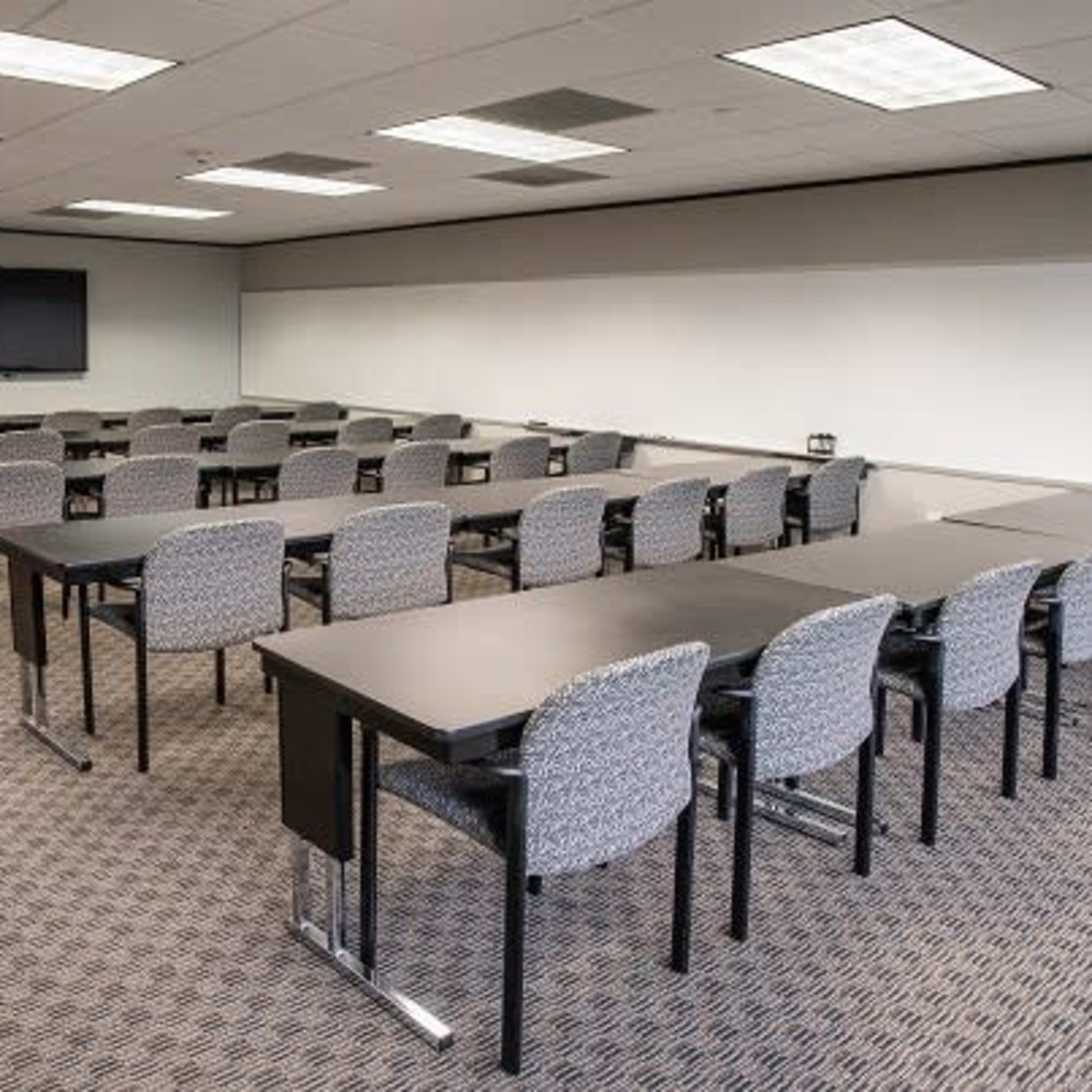 A spacious conference room with multiple rows of tables and chairs arranged for a meeting, featuring a large screen on one wall.