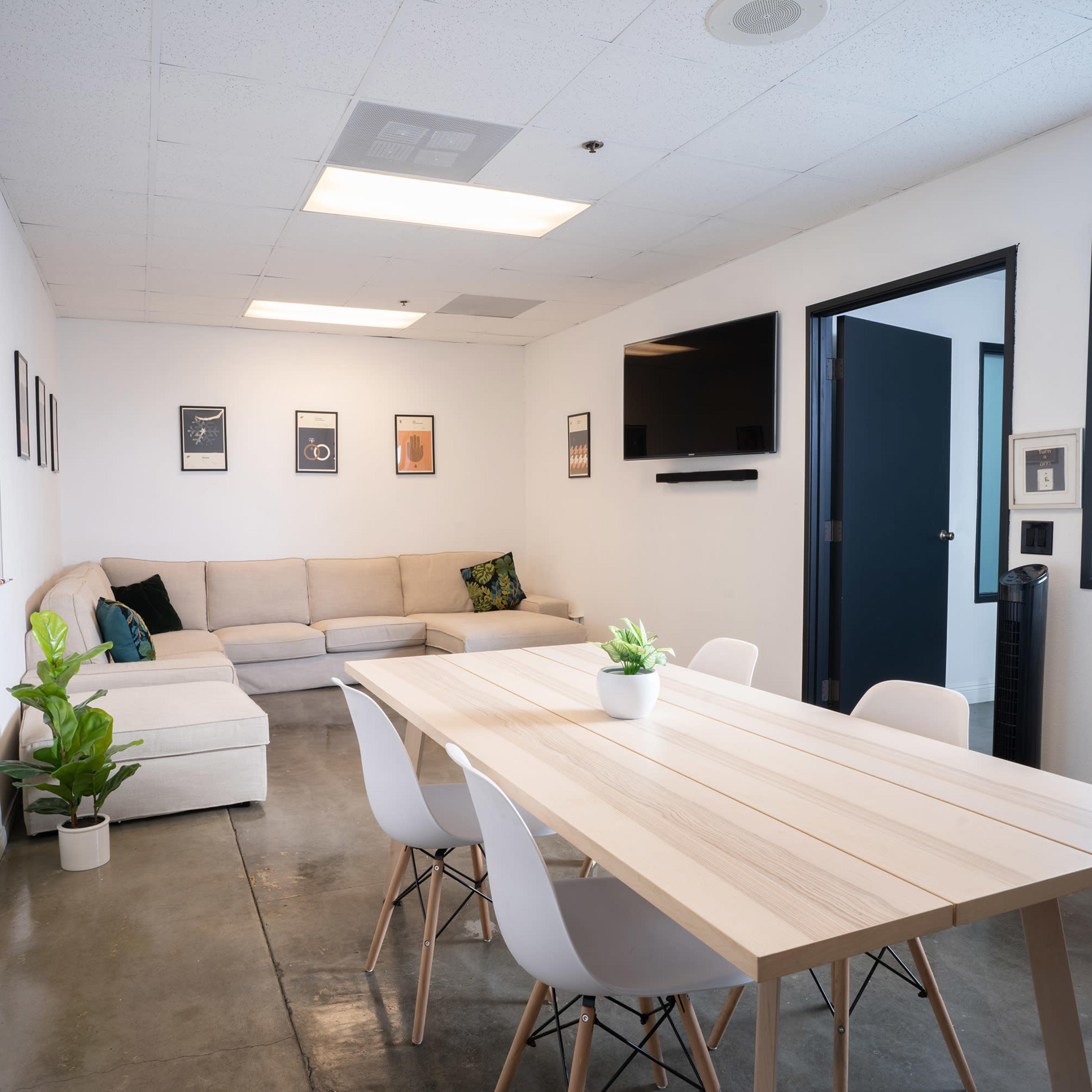 A modern meeting room with a sectional sofa, a wooden dining table surrounded by white chairs, and a television mounted on the wall.