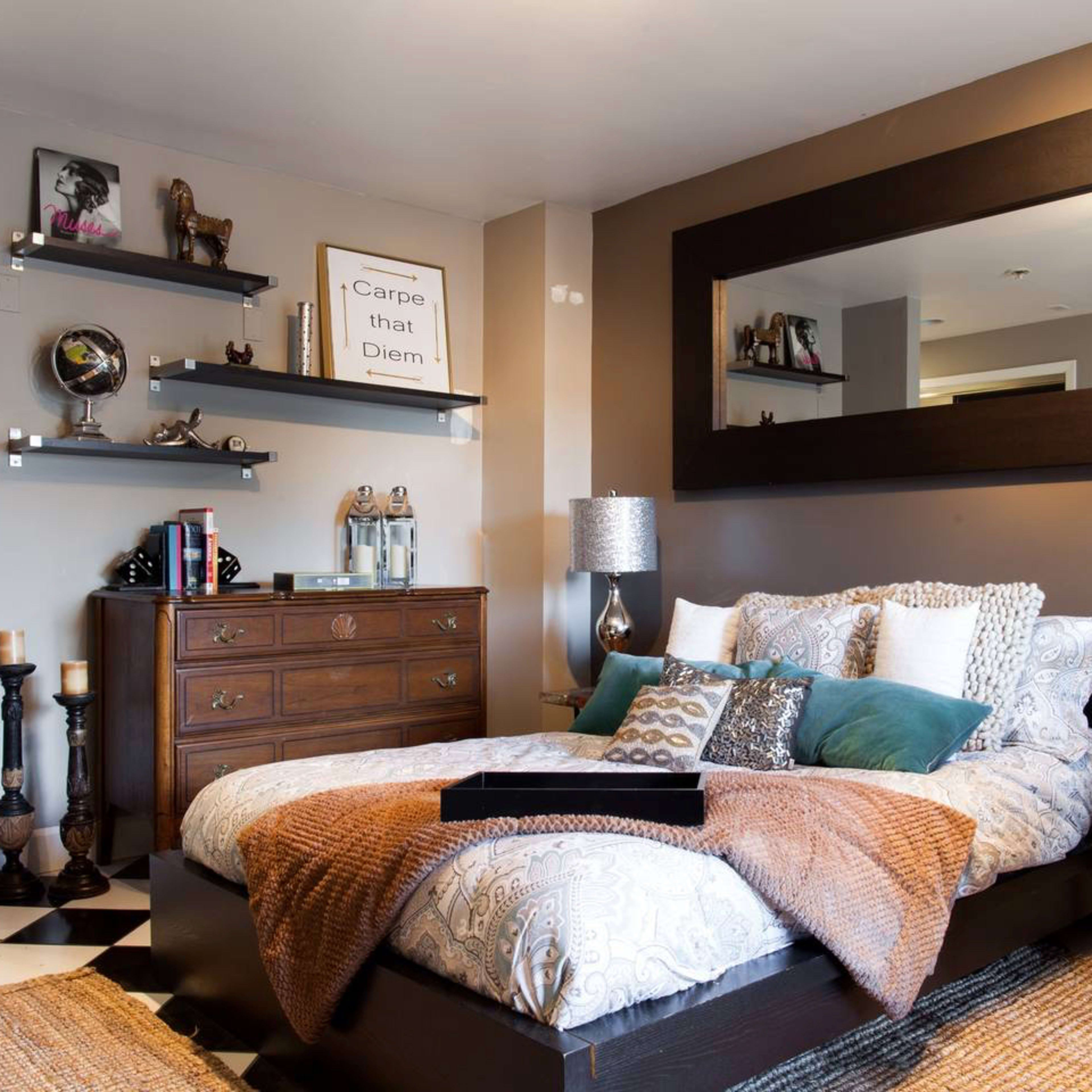 A cozy bedroom with a large bed adorned with various pillows and textured throws, a wooden dresser, and shelves displaying decorative items on a checkered floor.