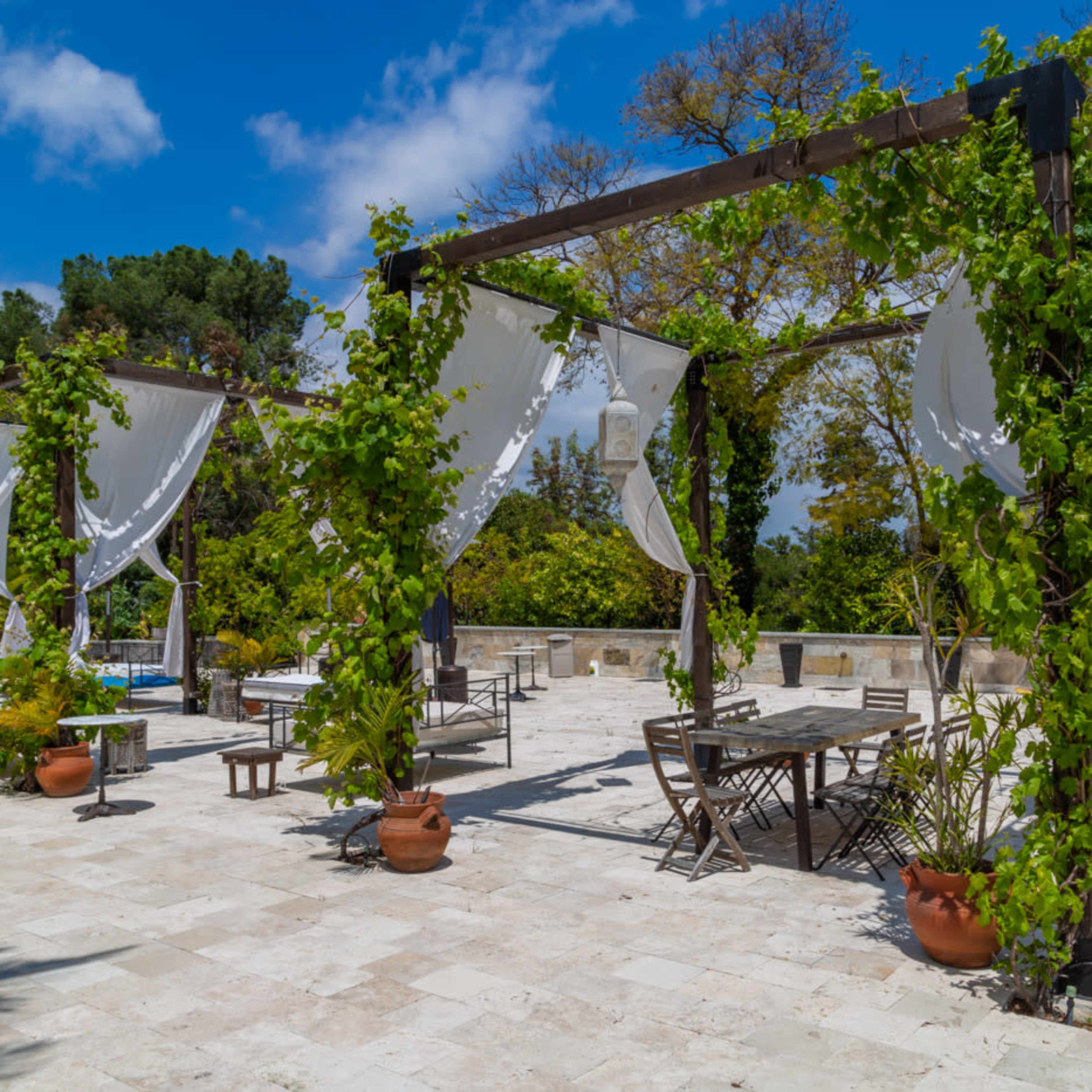 A stone terrace with wooden framework covered in climbing vines and draped fabric offers seating arrangements amidst a green landscape and blue sky.