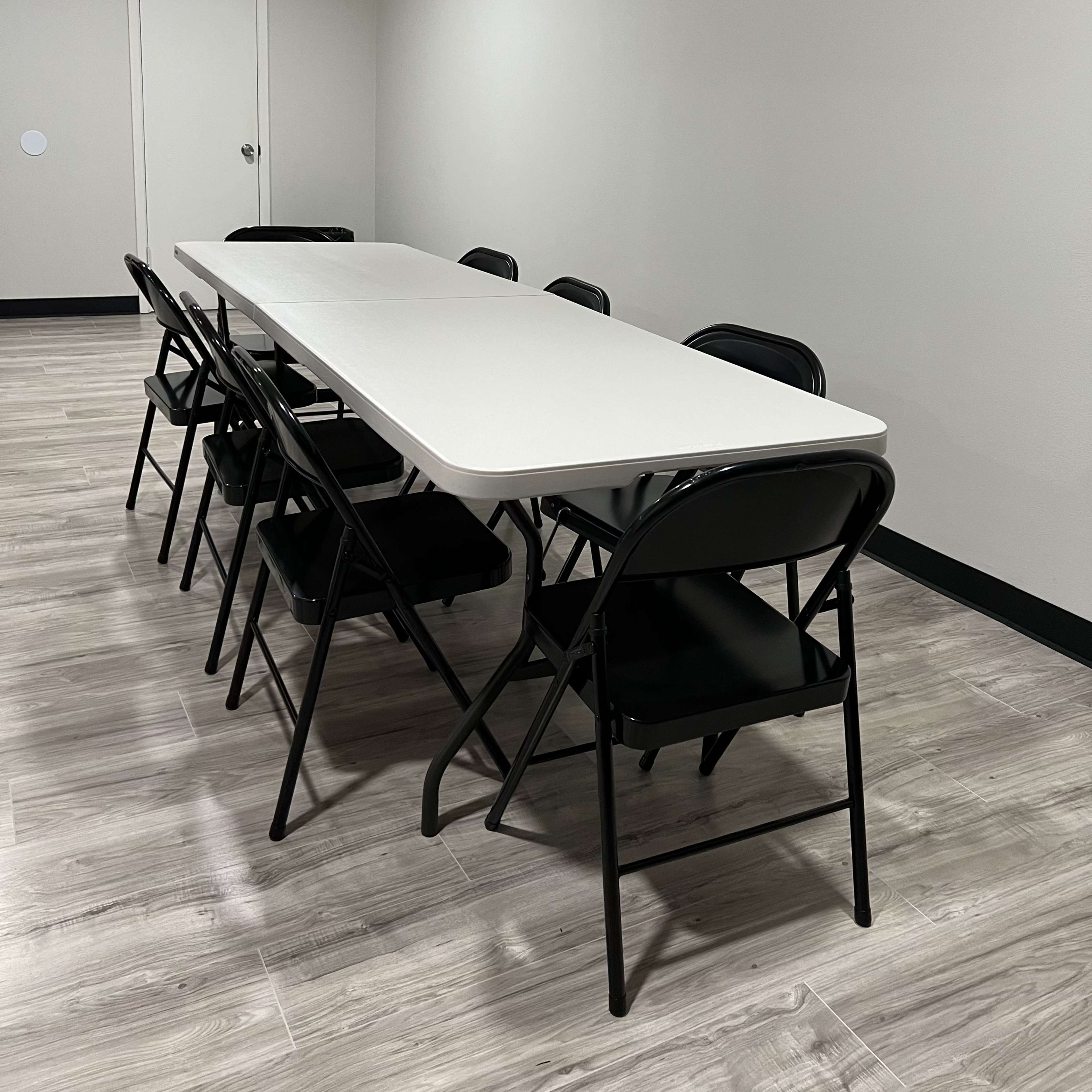 A long, rectangular white table is set in a brightly lit room with six black folding chairs arranged around it.