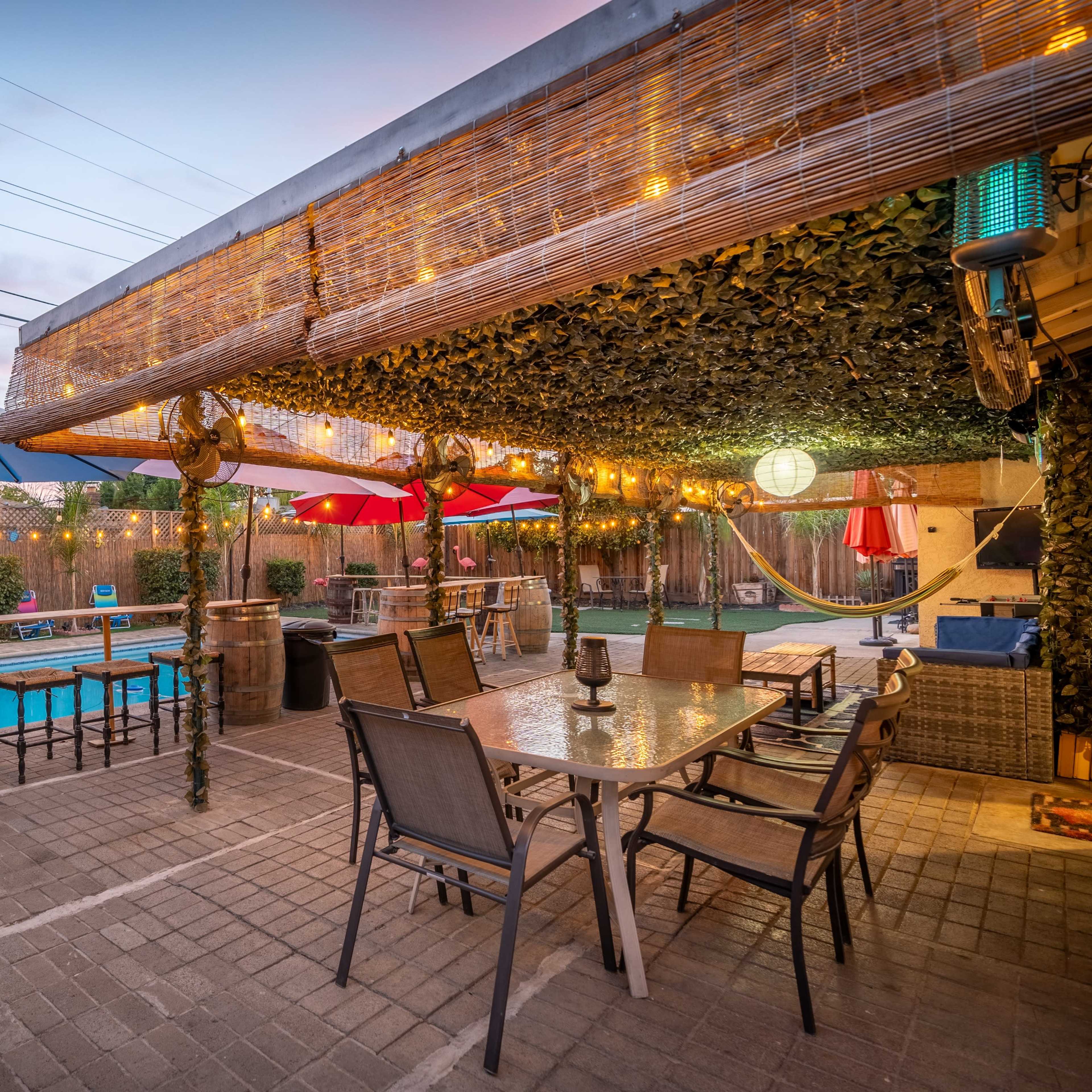 The outdoor space features a pool surrounded by seating areas, a dining table with chairs, and shaded umbrellas, all under decorative string lights.