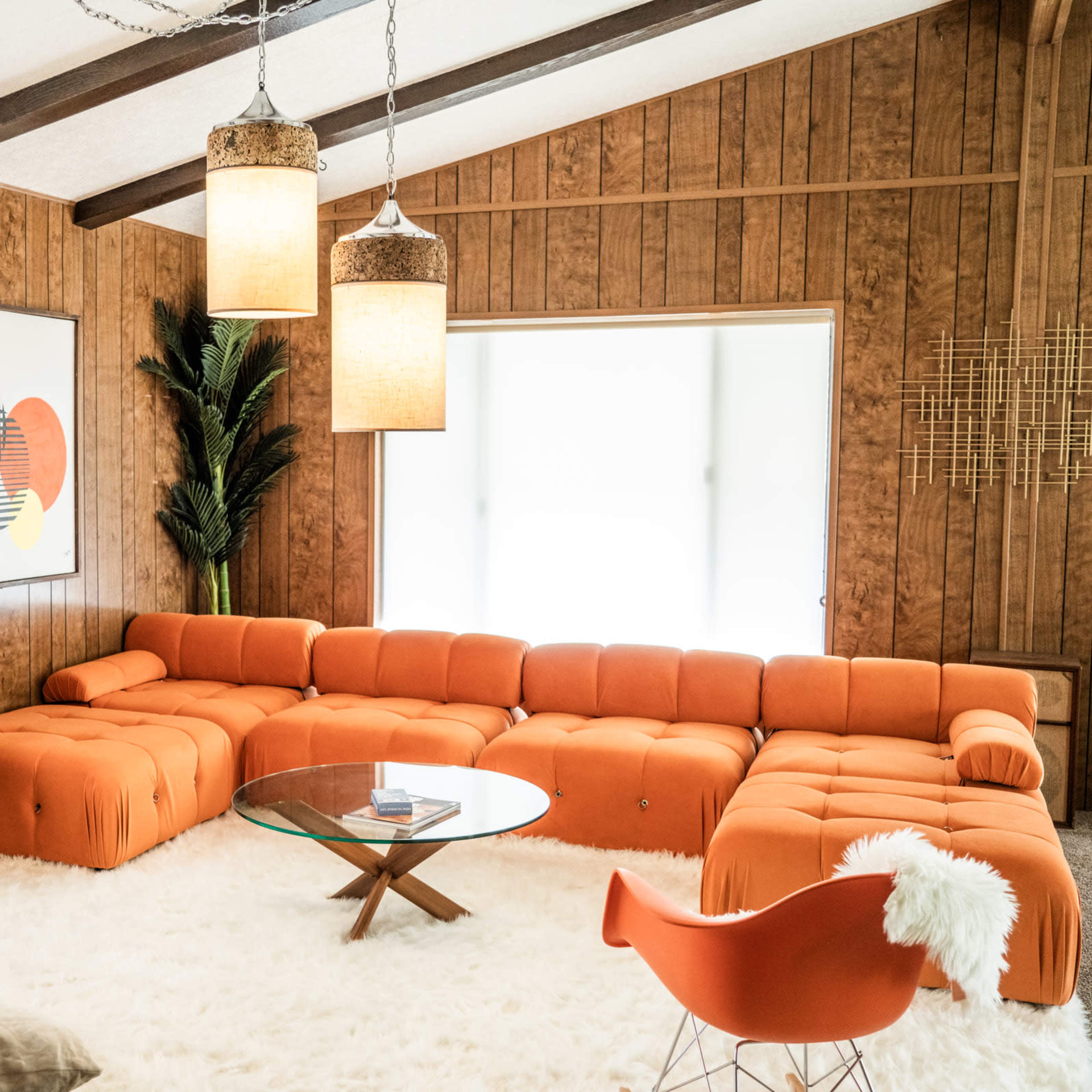 A modern living room with an orange sectional sofa, a glass coffee table, and wooden paneling on the walls.