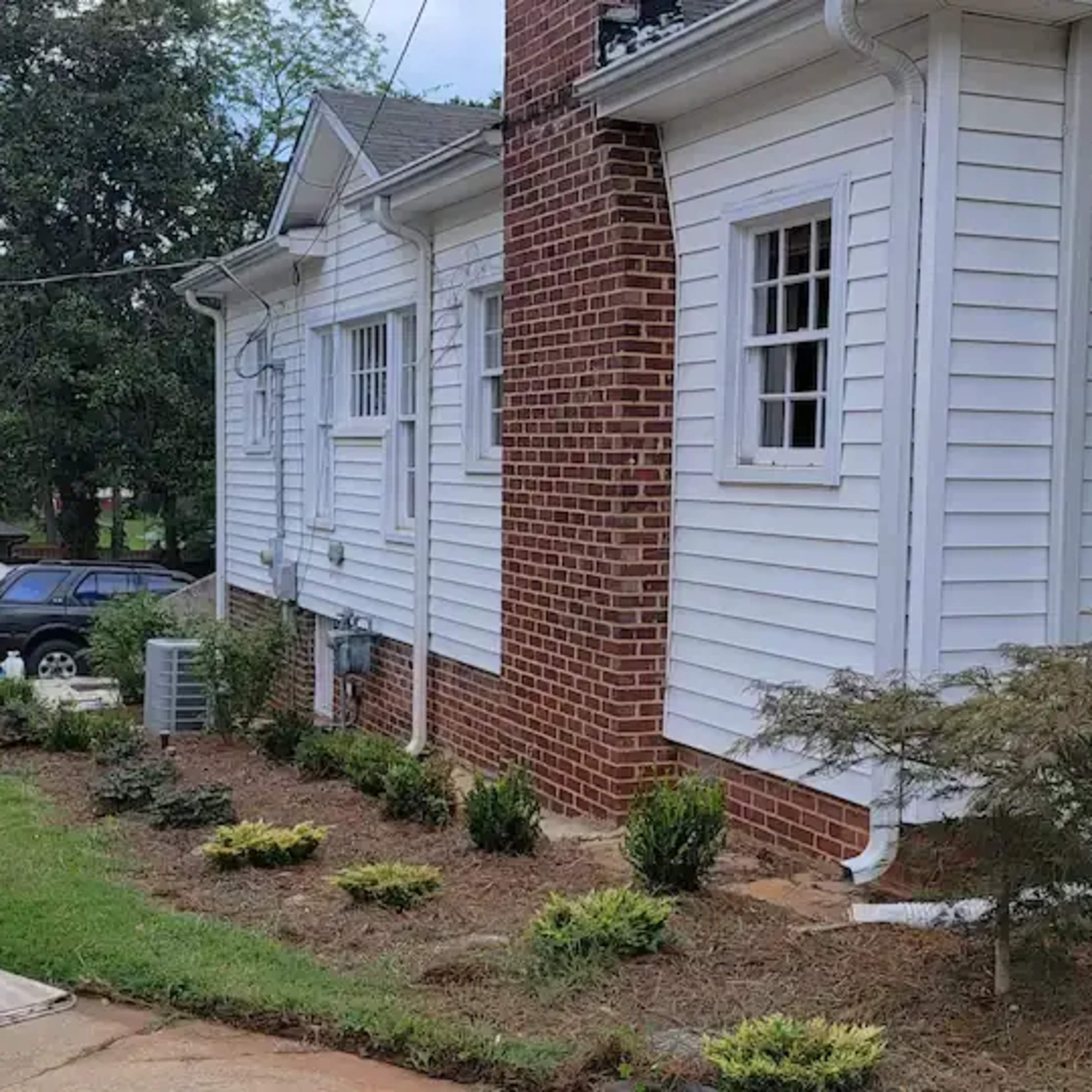 A white house with a brick chimney, a gravel driveway leading to a carport, and a garden area with shrubs and plants along the side.
