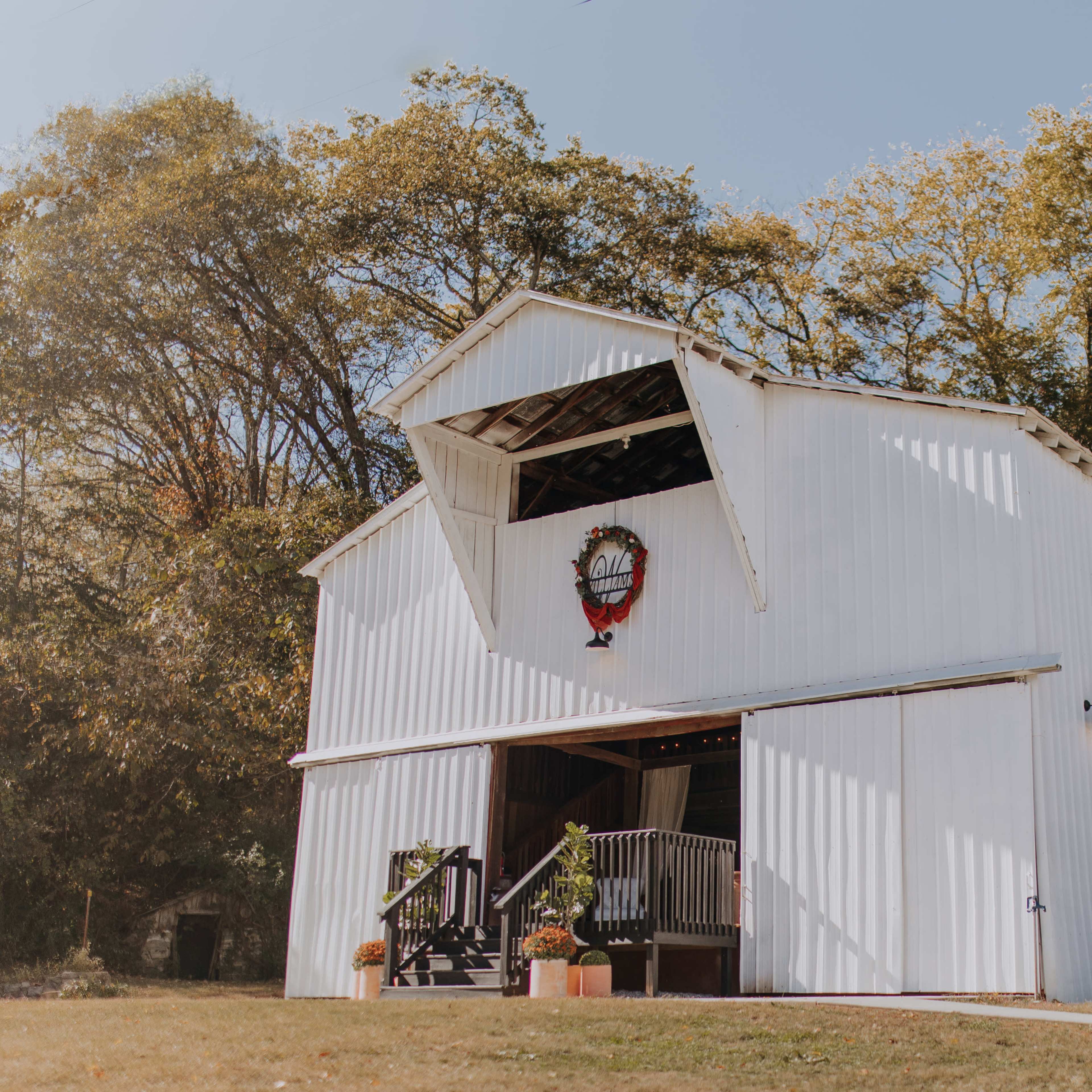A white barn with a large opening at the top and decorative elements stands on a grassy area surrounded by trees.