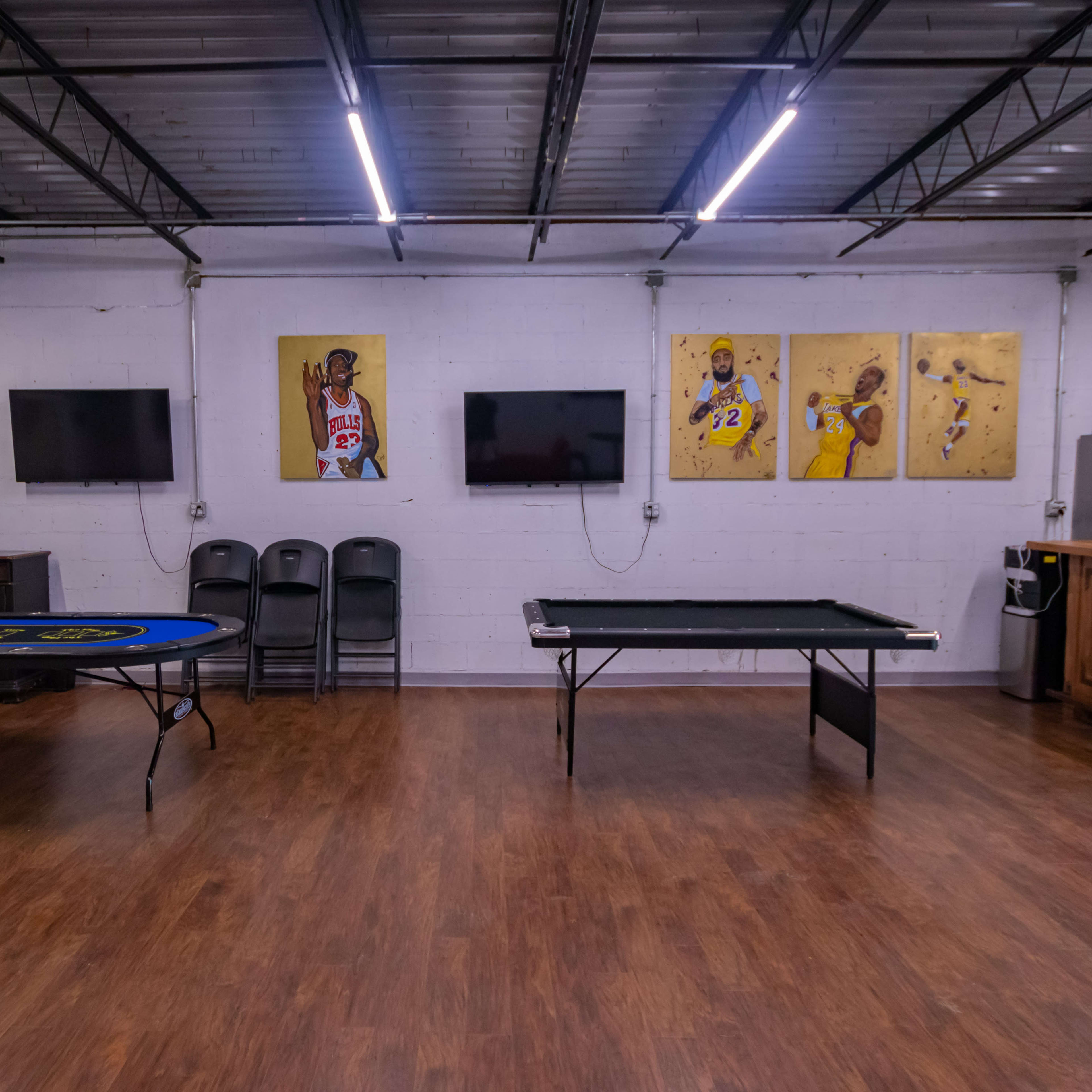 A recreational room with a blue poker table on the left, a black pool table on the right, and two mounted televisions along with framed artwork of basketball players on the walls.