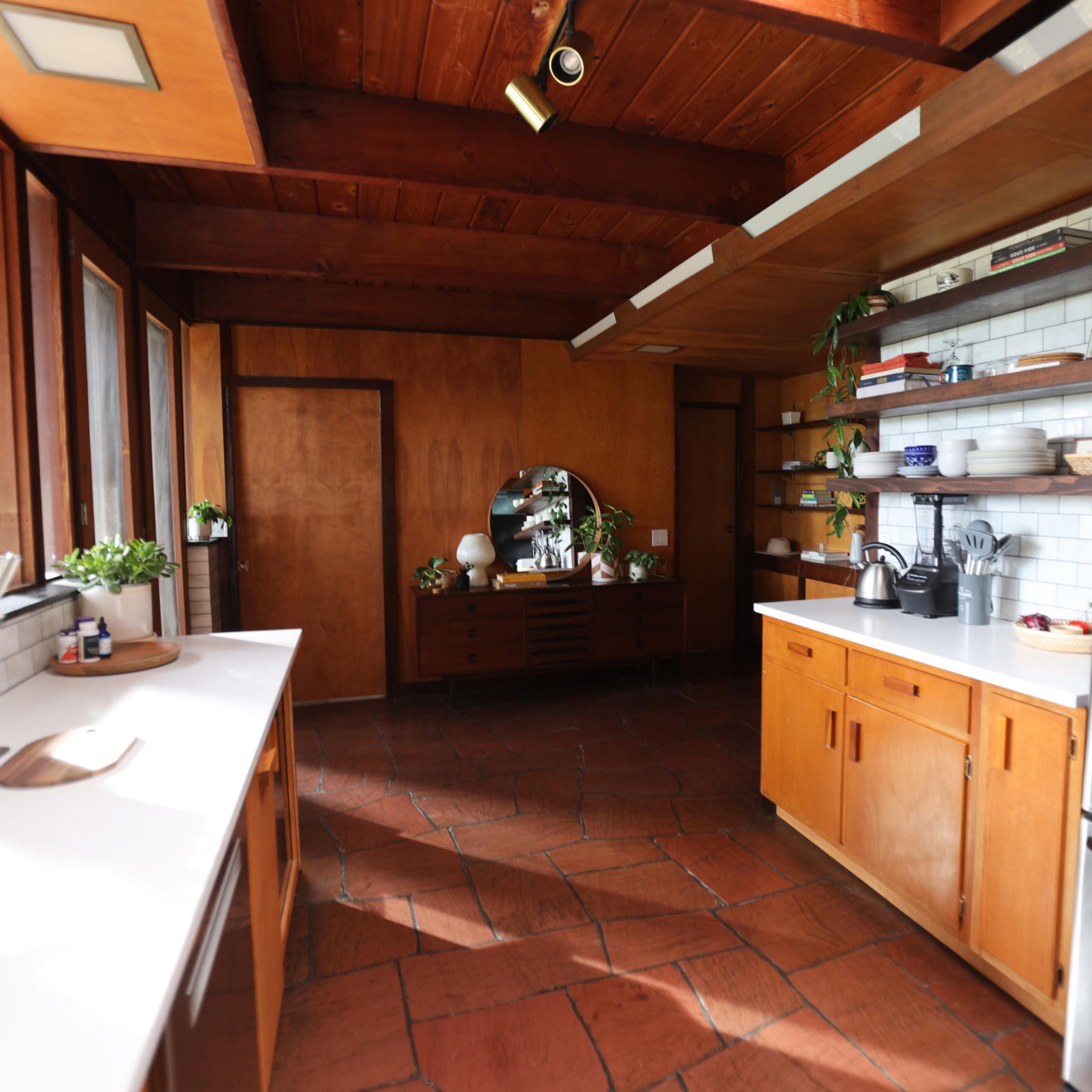 A modern kitchen featuring wooden cabinets, open shelving with kitchenware, and stainless steel appliances, all set against a backdrop of terracotta tile flooring.