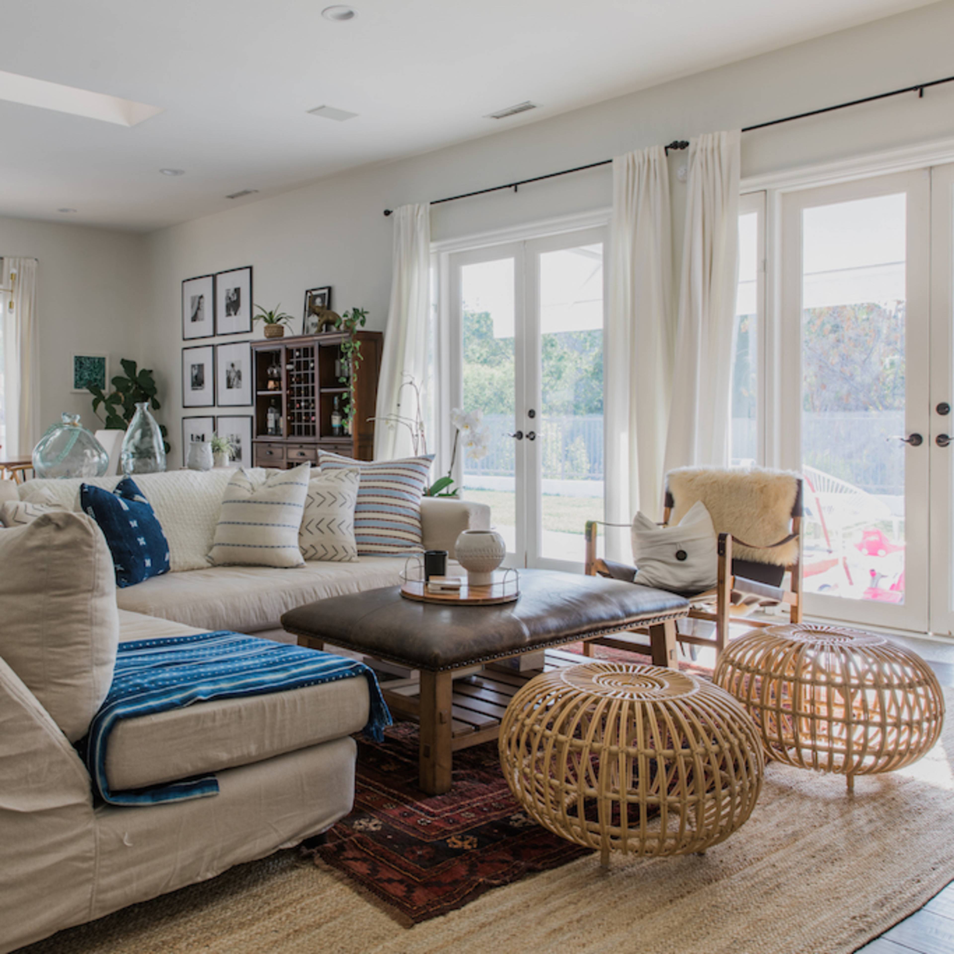 A spacious living room features a large beige sectional sofa, wooden coffee table, and decorative woven poufs, with natural light streaming in through sliding glass doors.