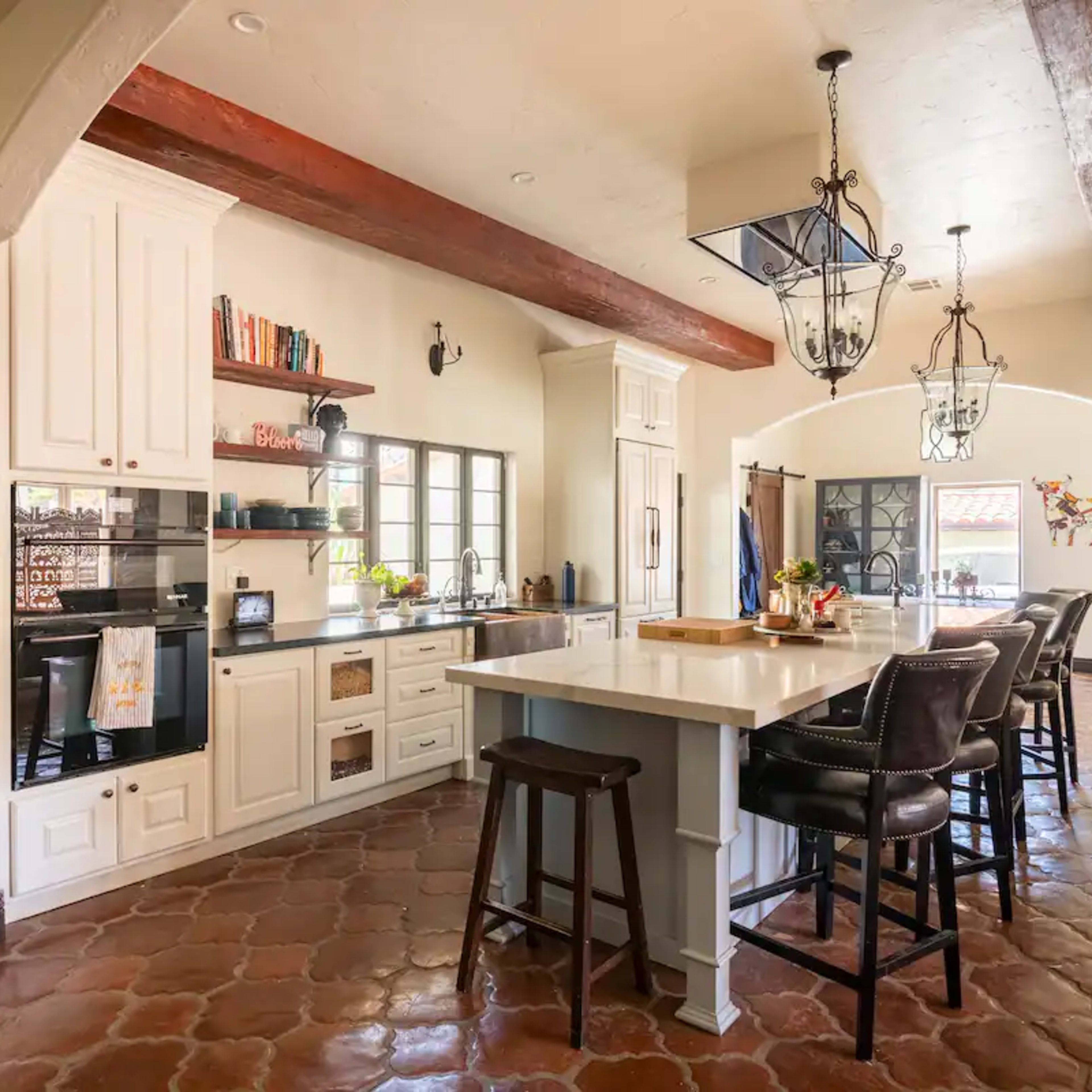 A bright, spacious kitchen with white cabinetry, a large island, and a mix of tile and wooden flooring, complemented by a dining area featuring tall chairs.