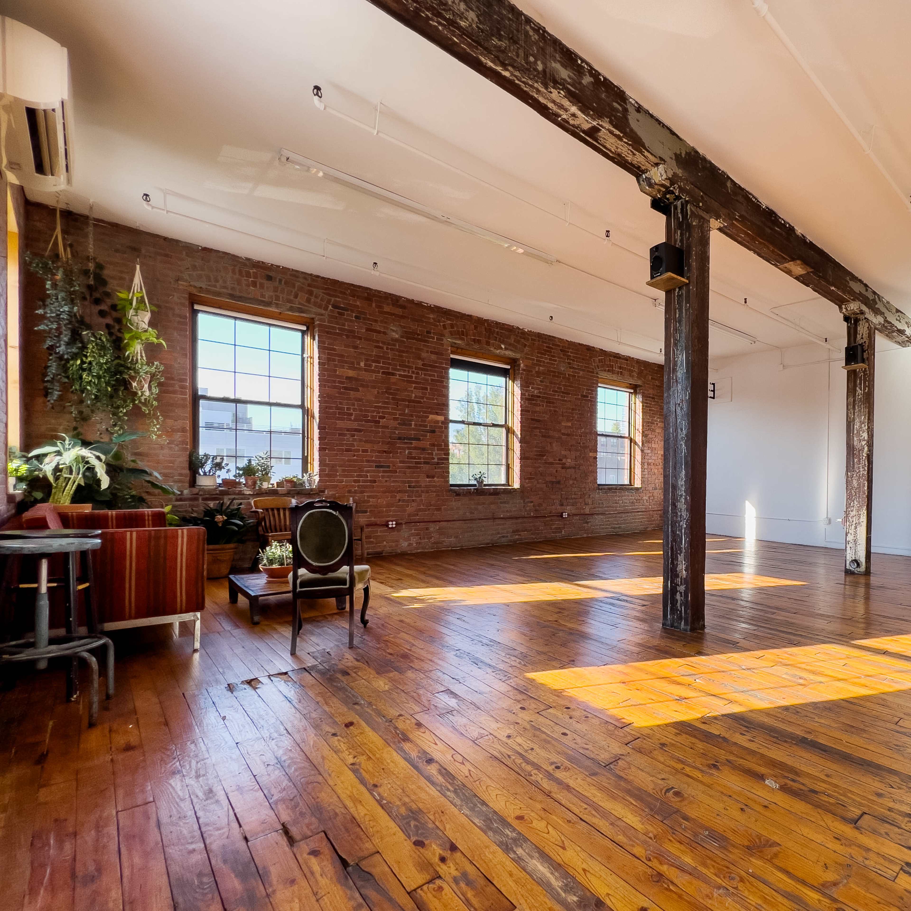 A spacious, well-lit room with hardwood floors, large windows, exposed brick walls, and wooden beams.