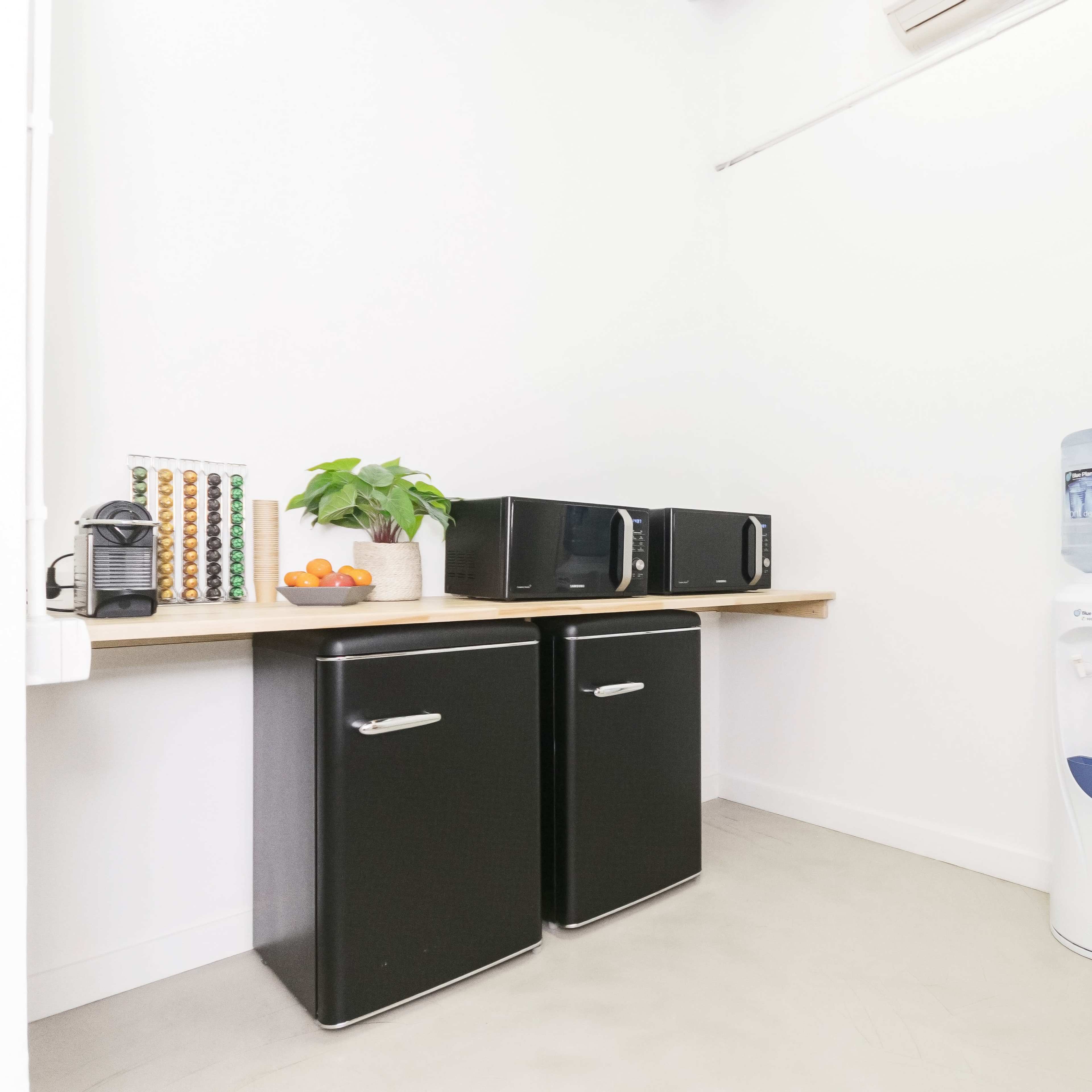 A small kitchen space featuring two black refrigerators, a countertop with a plant and fruit bowl, and a water cooler against the wall.