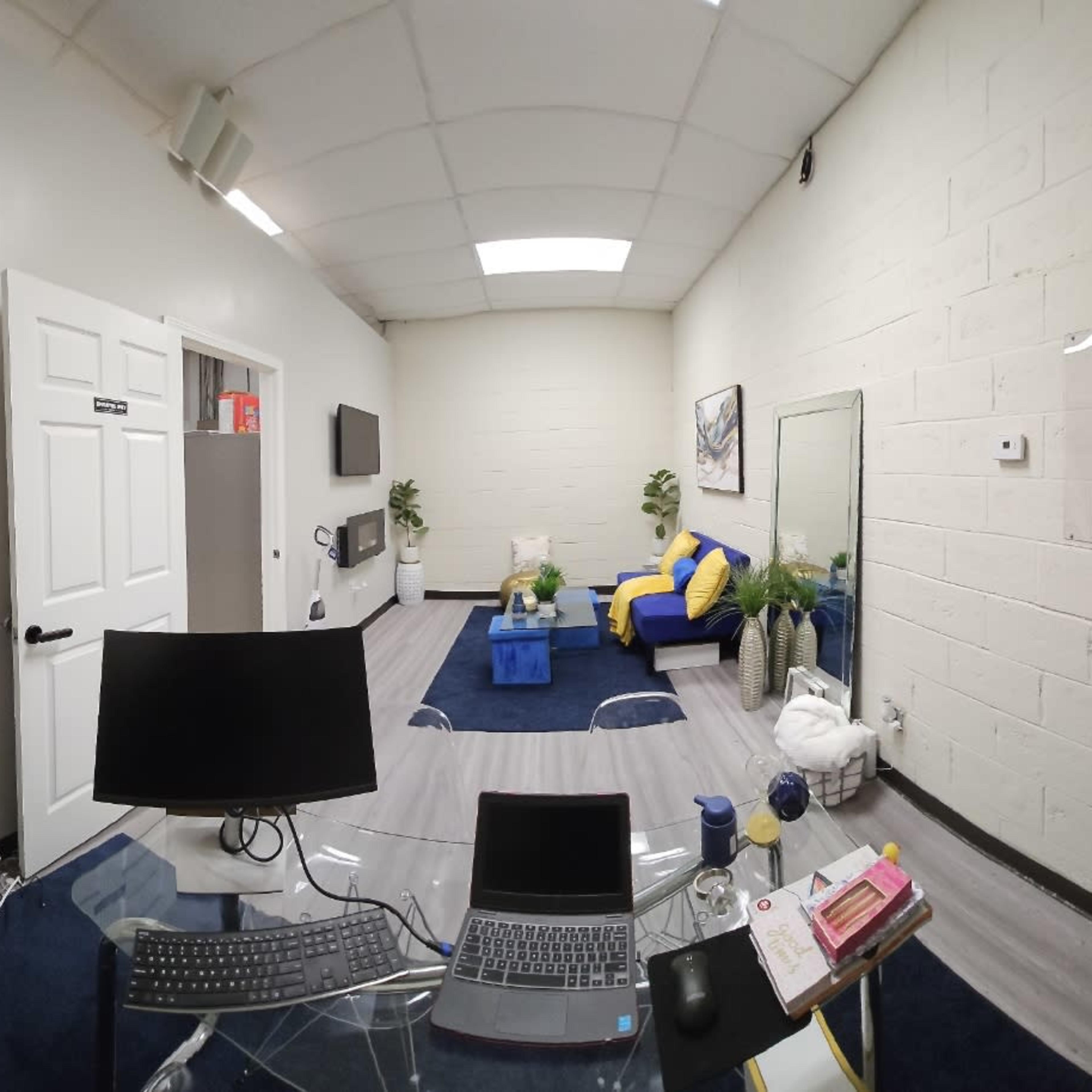 A small office space featuring a desk with a laptop, a couch, a blue table, and decorative plants on the floor.
