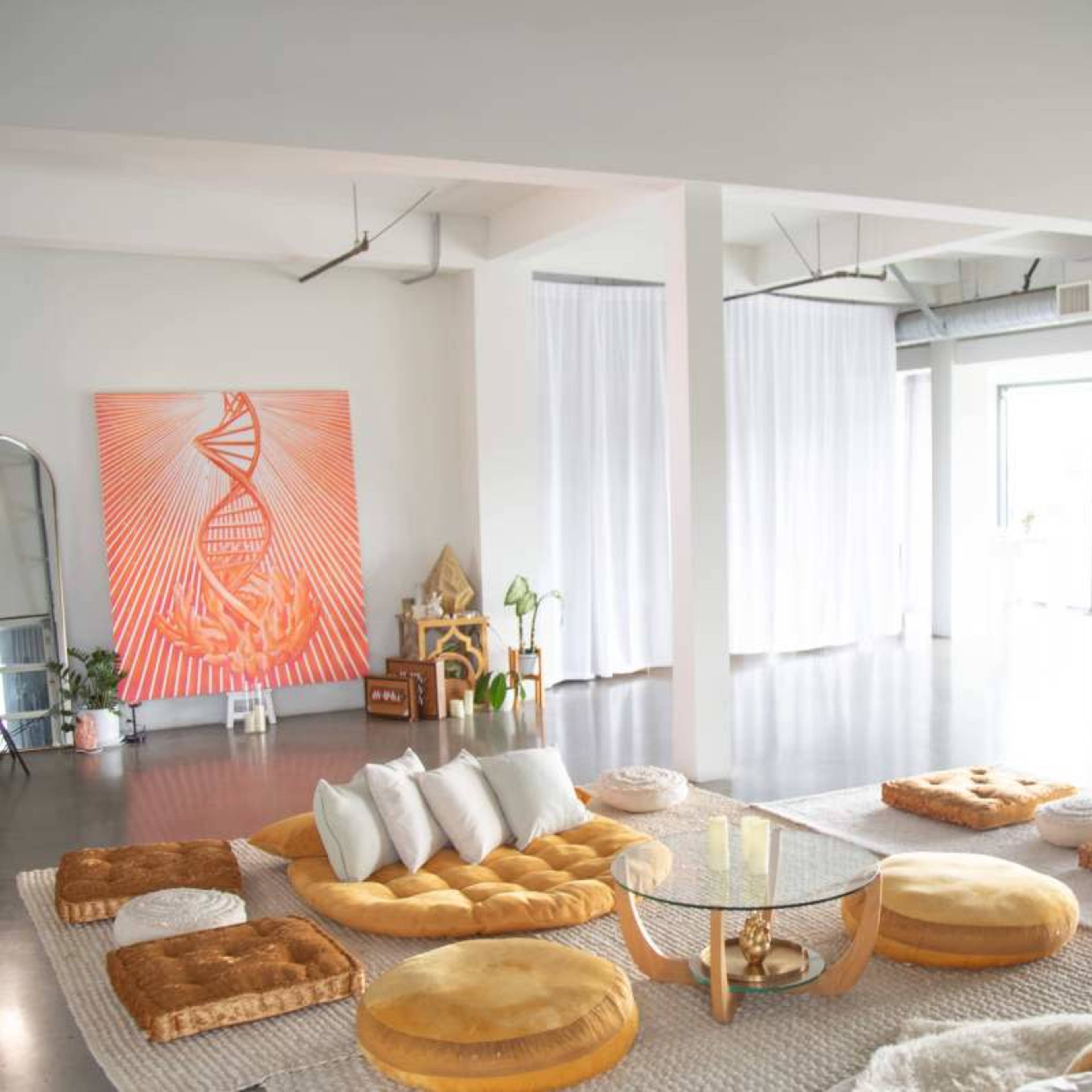 A spacious, modern room filled with various cushions and seating arrangements, large windows letting in natural light, and a vibrant artwork on the wall.