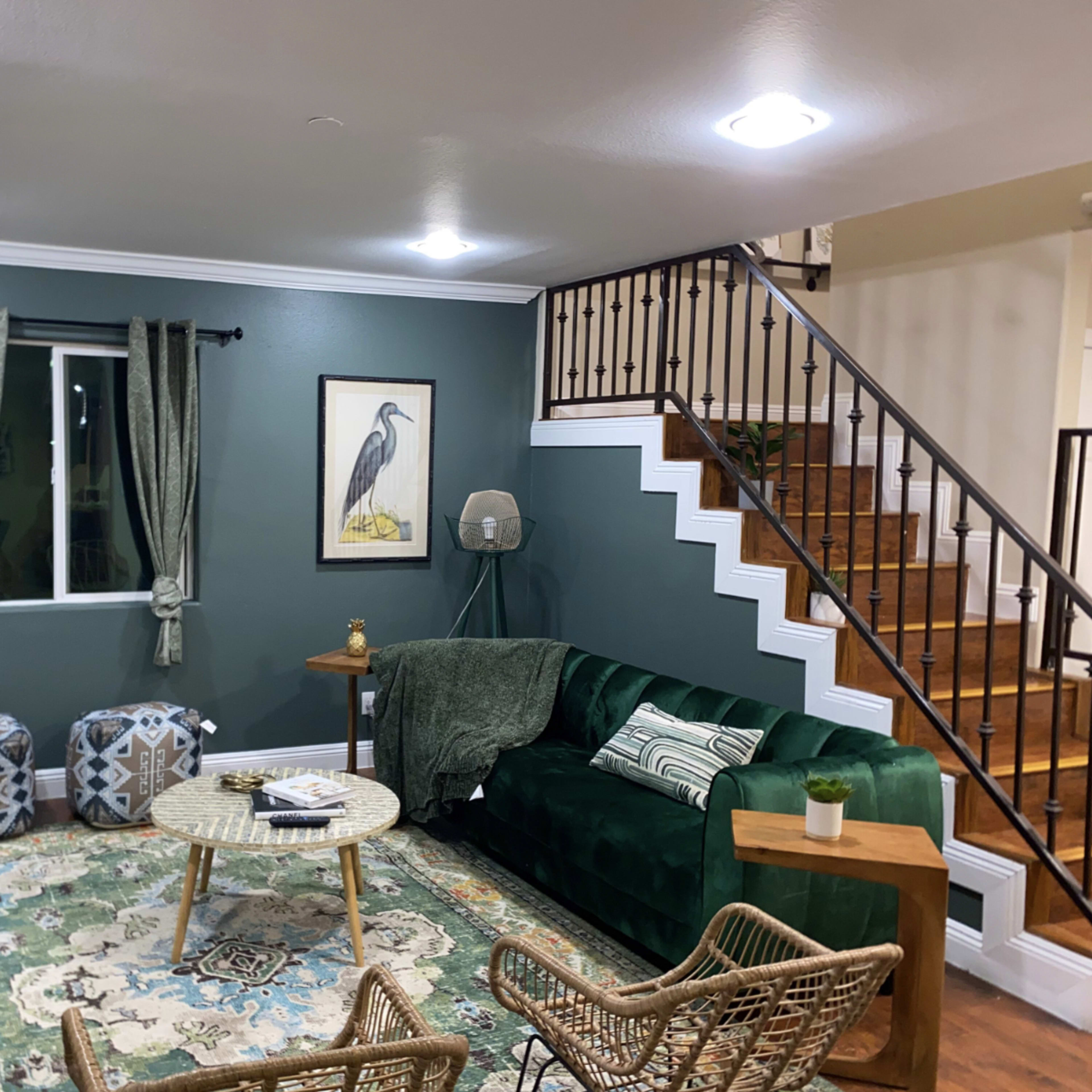 A living room featuring a green sofa, decorative poufs, a round mirror, and a staircase with wooden steps.