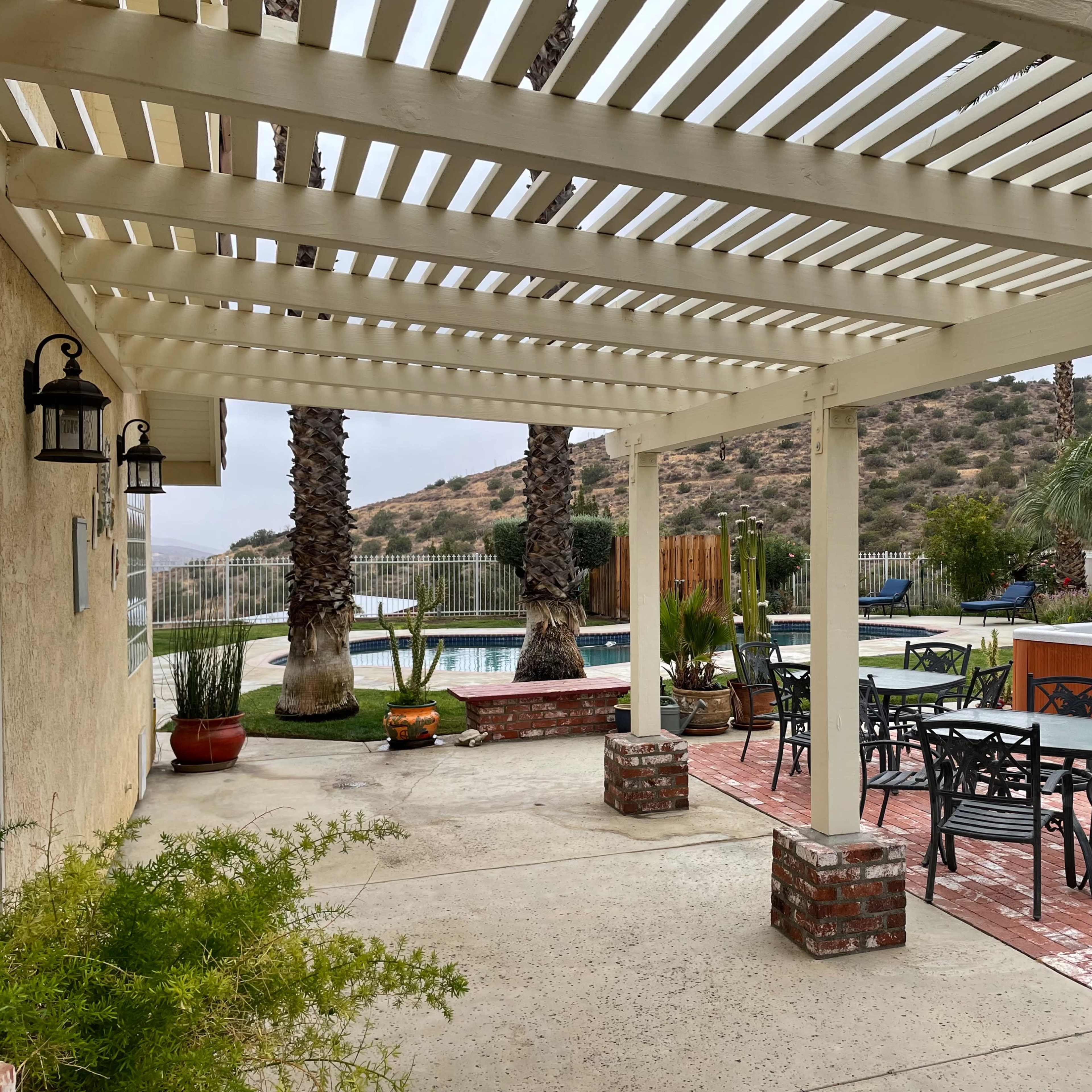 A patio area with a covered structure, tables, and chairs, overlooking a pool and landscaped hills in the background.