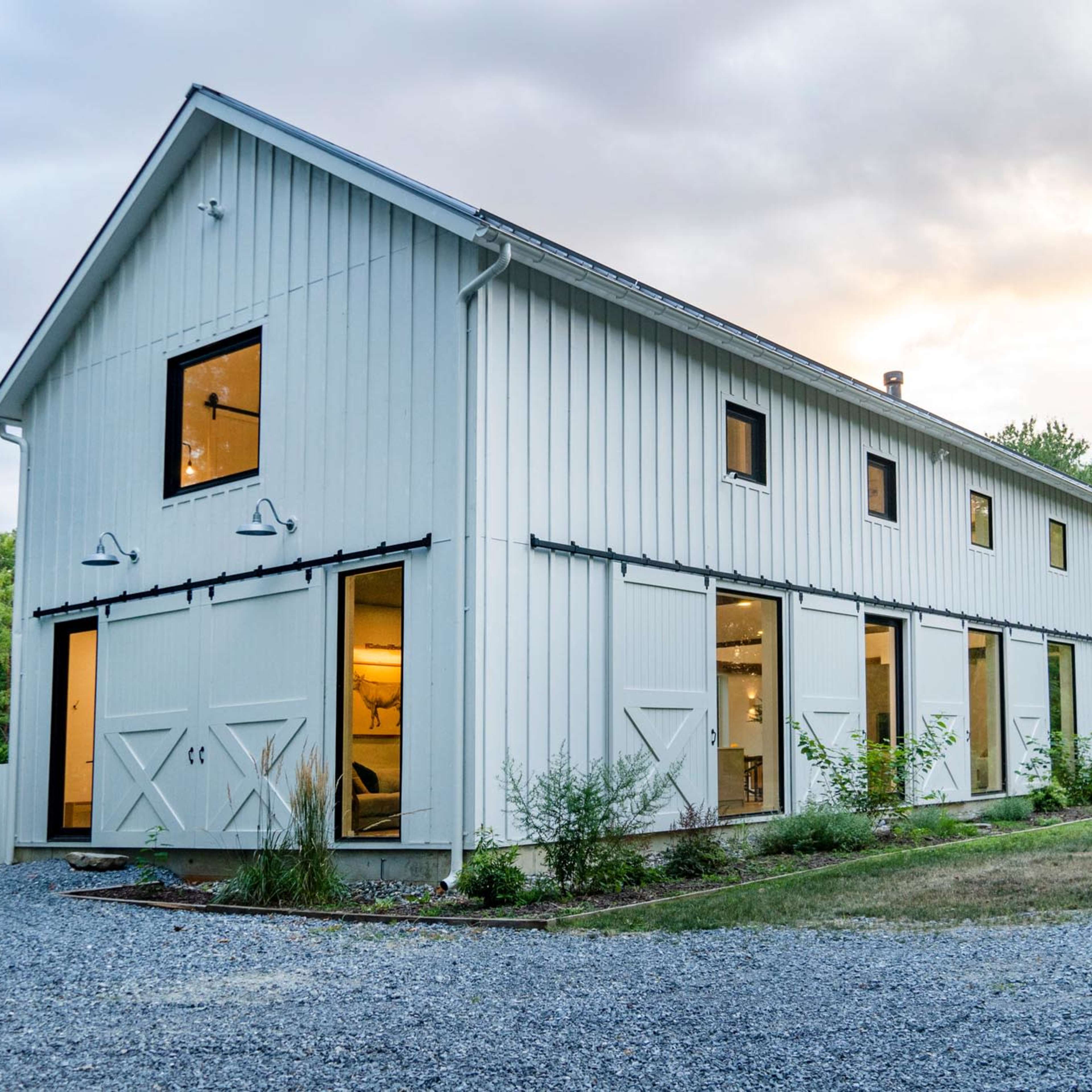 A white, two-story barn-style building with large windows and sliding doors is set on a gravel landscape surrounded by greenery.