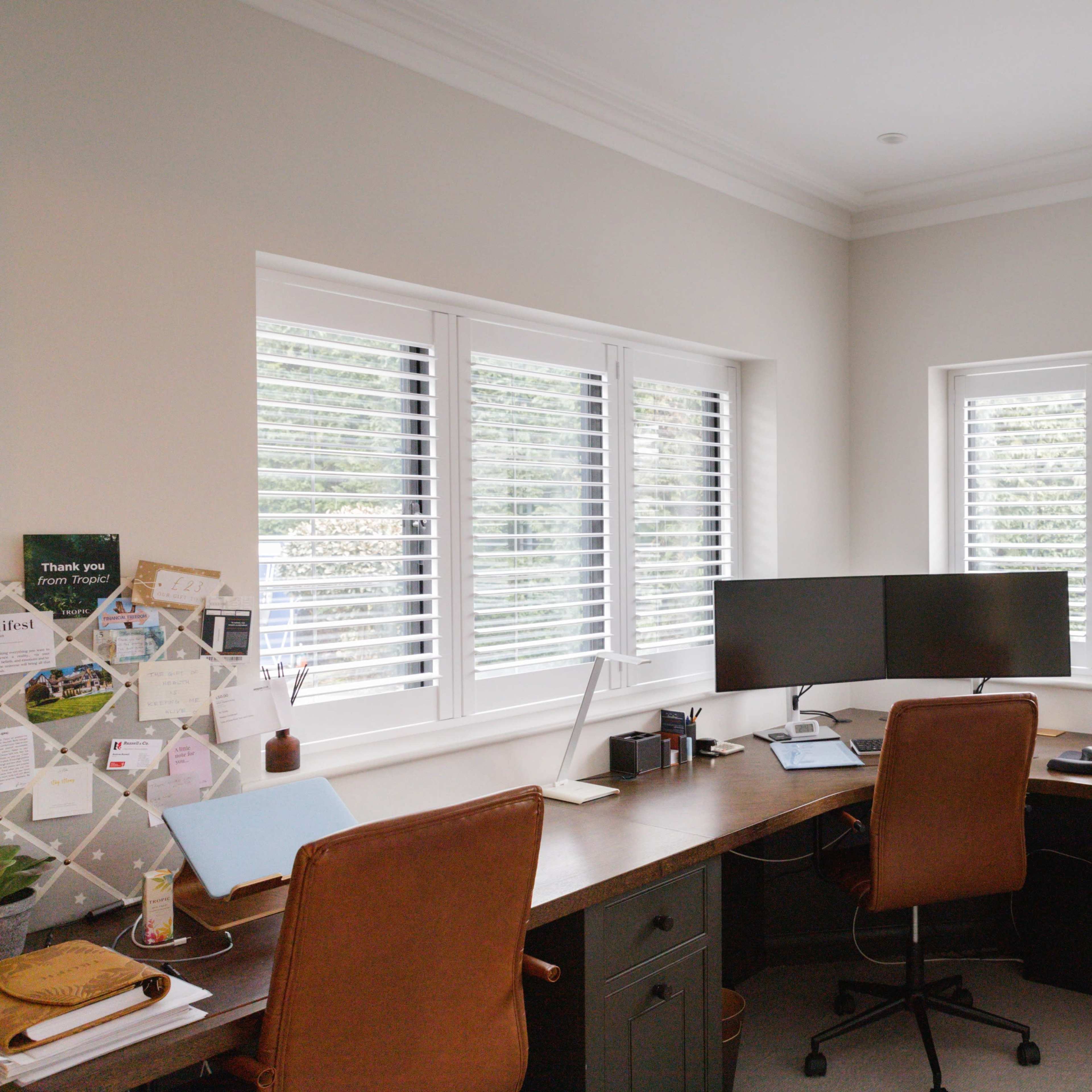A modern home office features a corner desk with a computer monitor and two leather chairs, surrounded by large windows with white shutters.