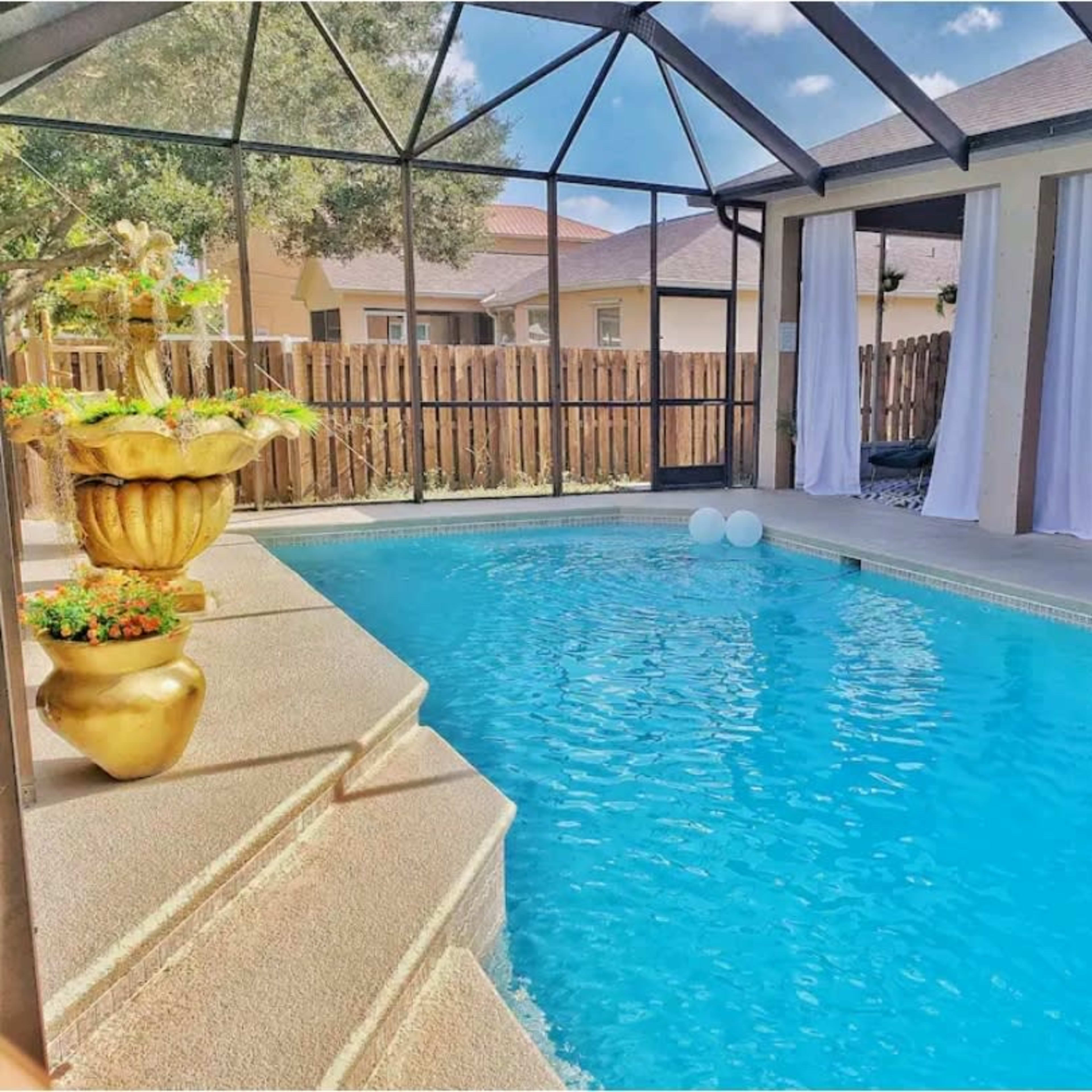 A screened-in pool area featuring a clear blue pool, a decorative planter, and seating arrangements with curtains on either side.