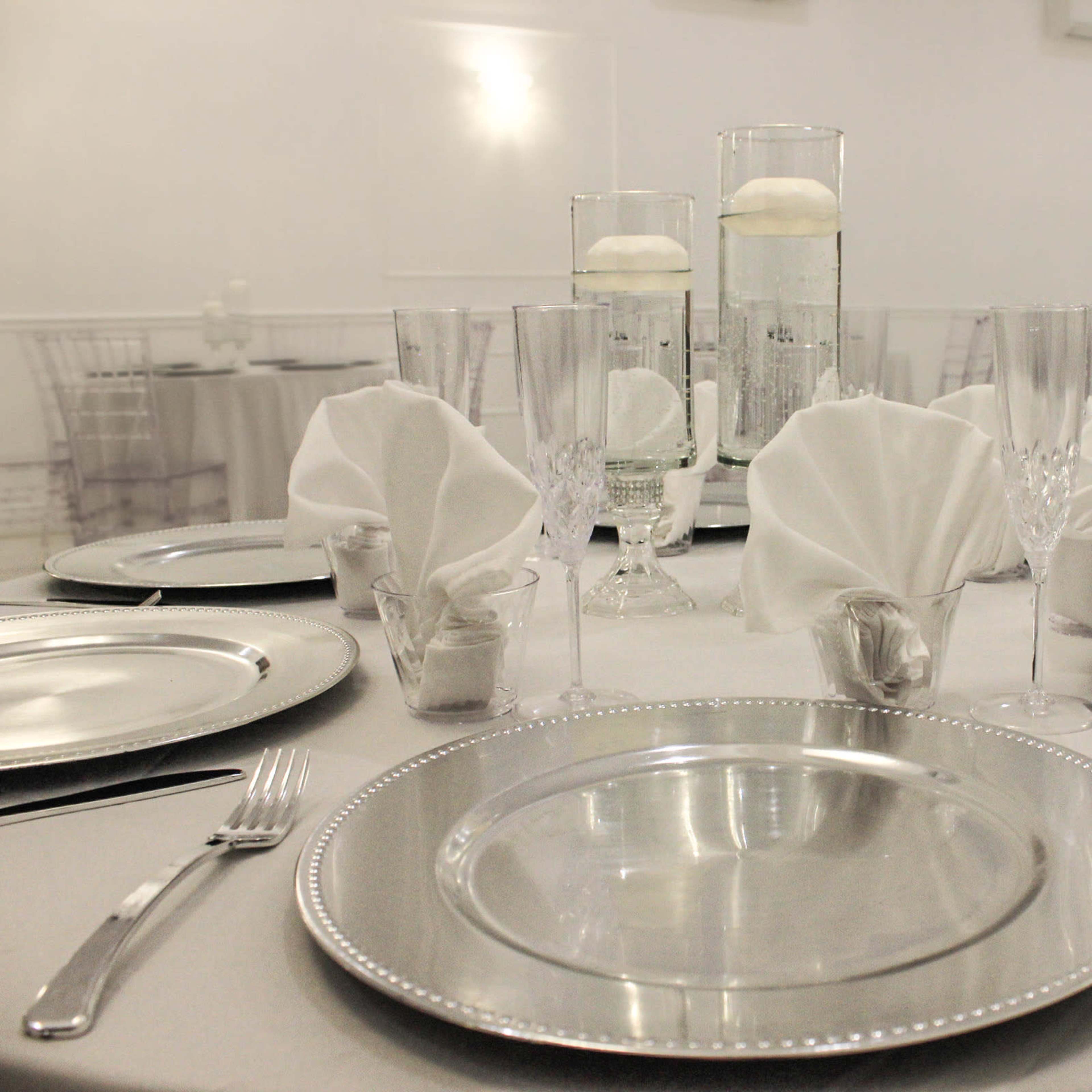 An elegantly arranged dining table set with silver chargers, crystal glassware, and folded napkins, with decorative centerpieces in clear vases.