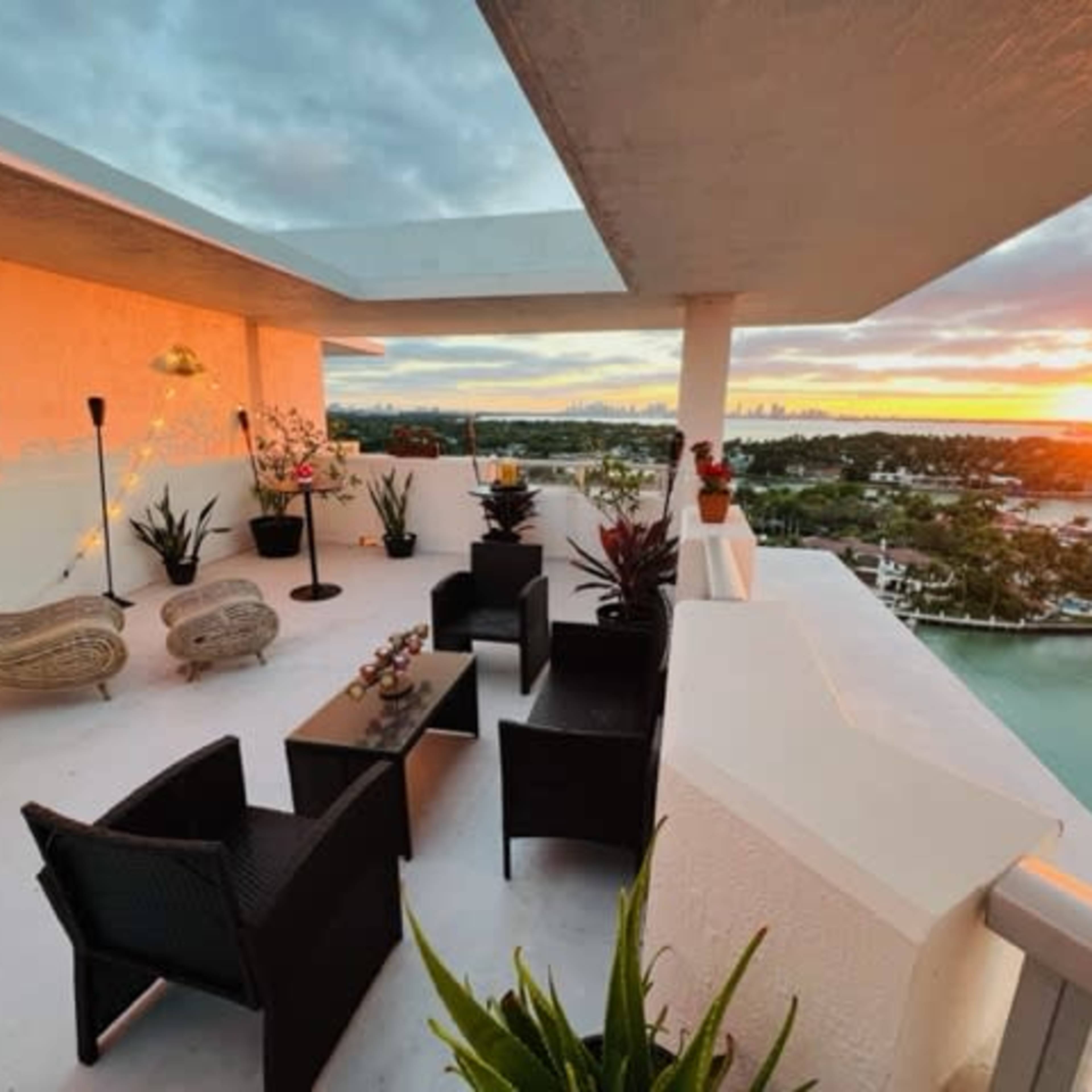 A terrace features modern seating and decorative plants overlooking a waterway at sunset.