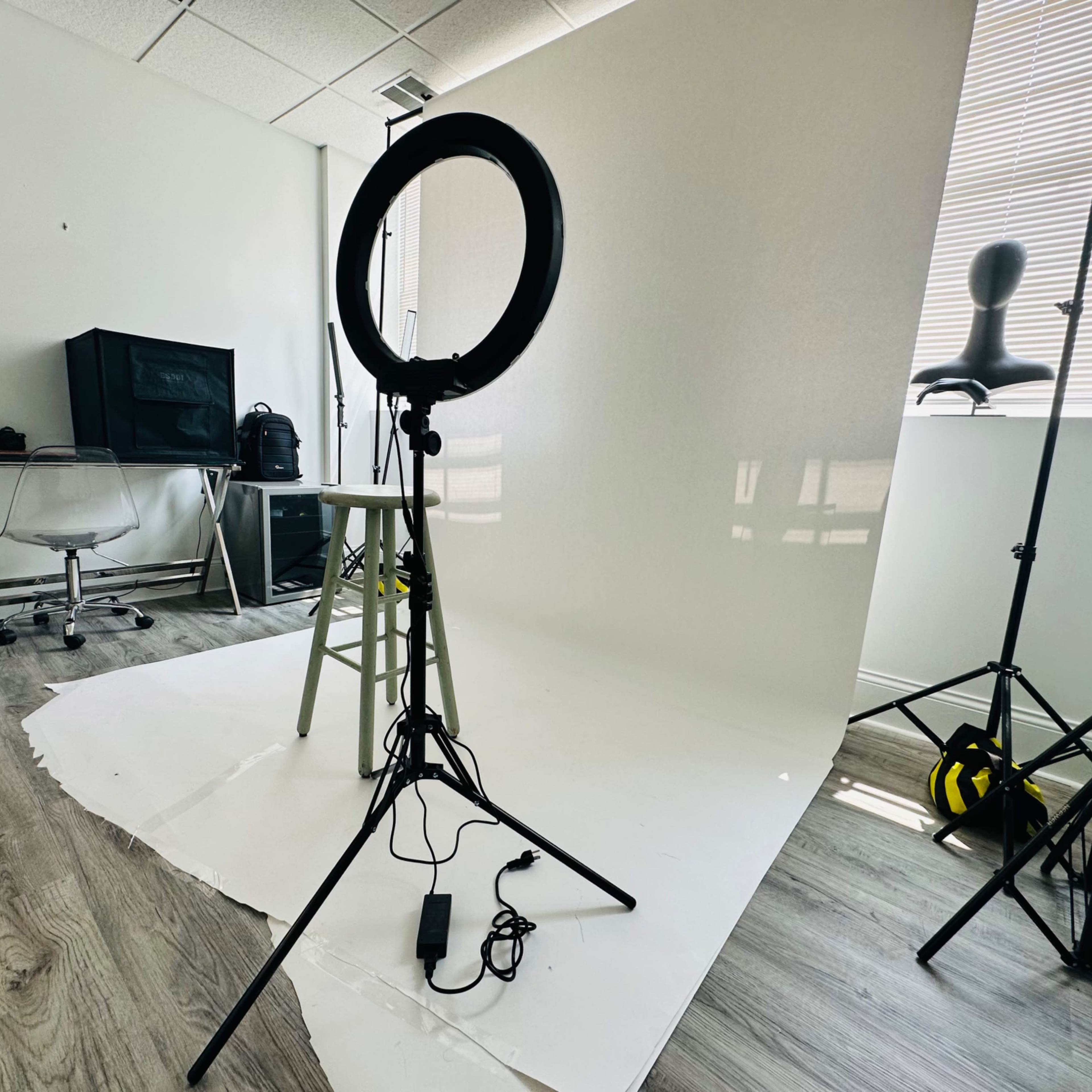 A photography studio setup featuring a ring light, a stool, and a white backdrop, with additional equipment and furniture in the background.