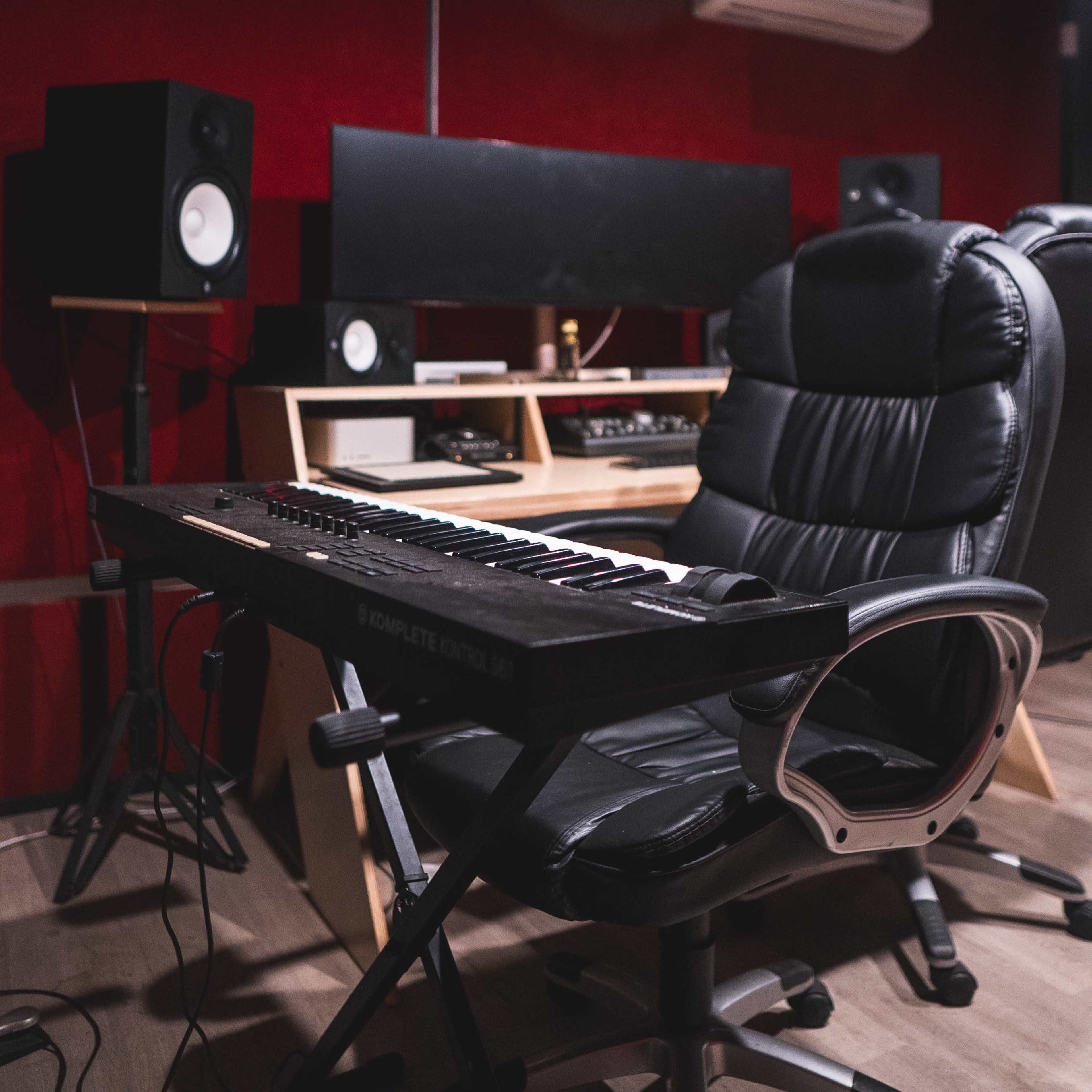A black keyboard sits on a folding chair in a music studio with speakers and a mixing console visible in the background.