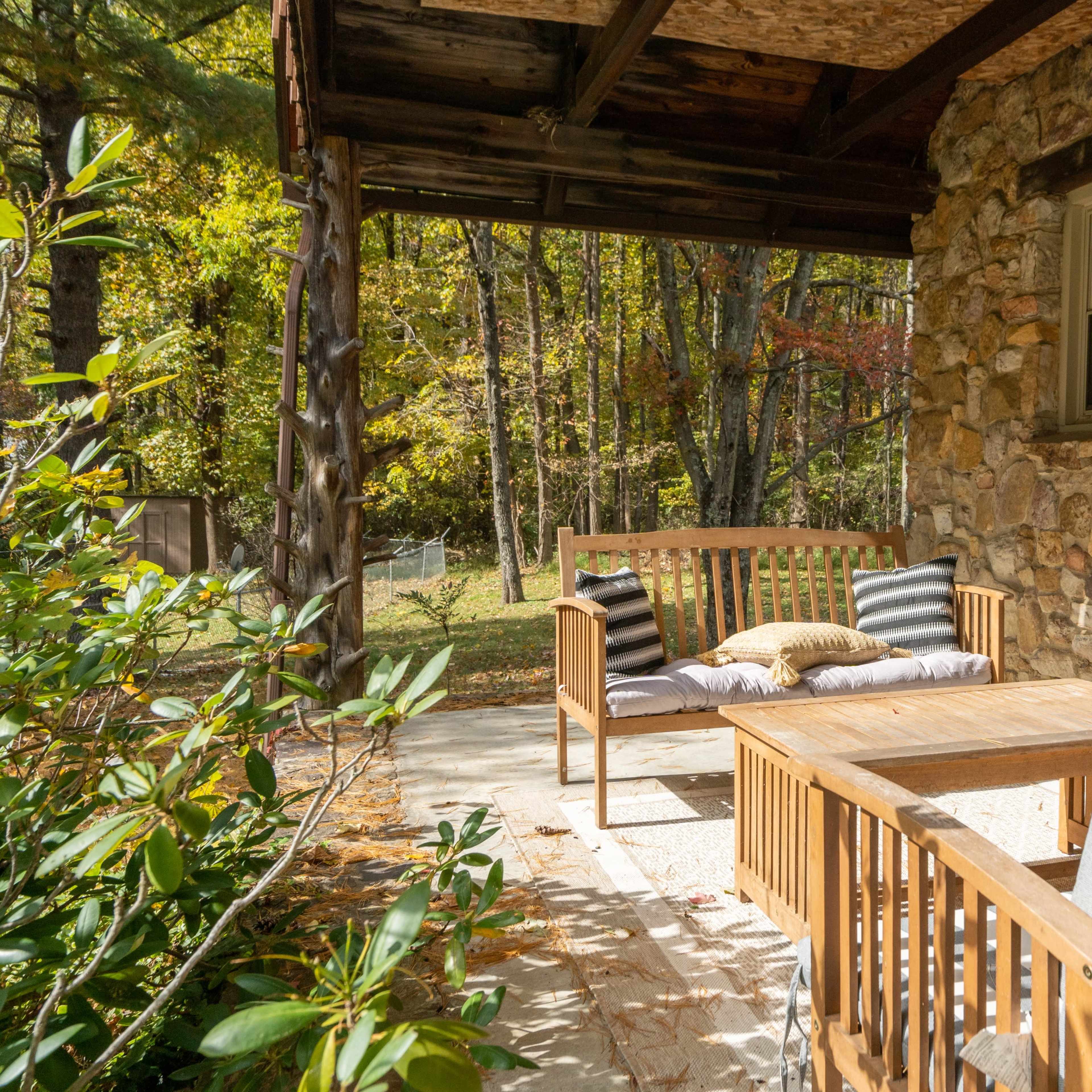 A wooden seating area with a table is positioned on a patio beside a stone wall, surrounded by greenery and trees.