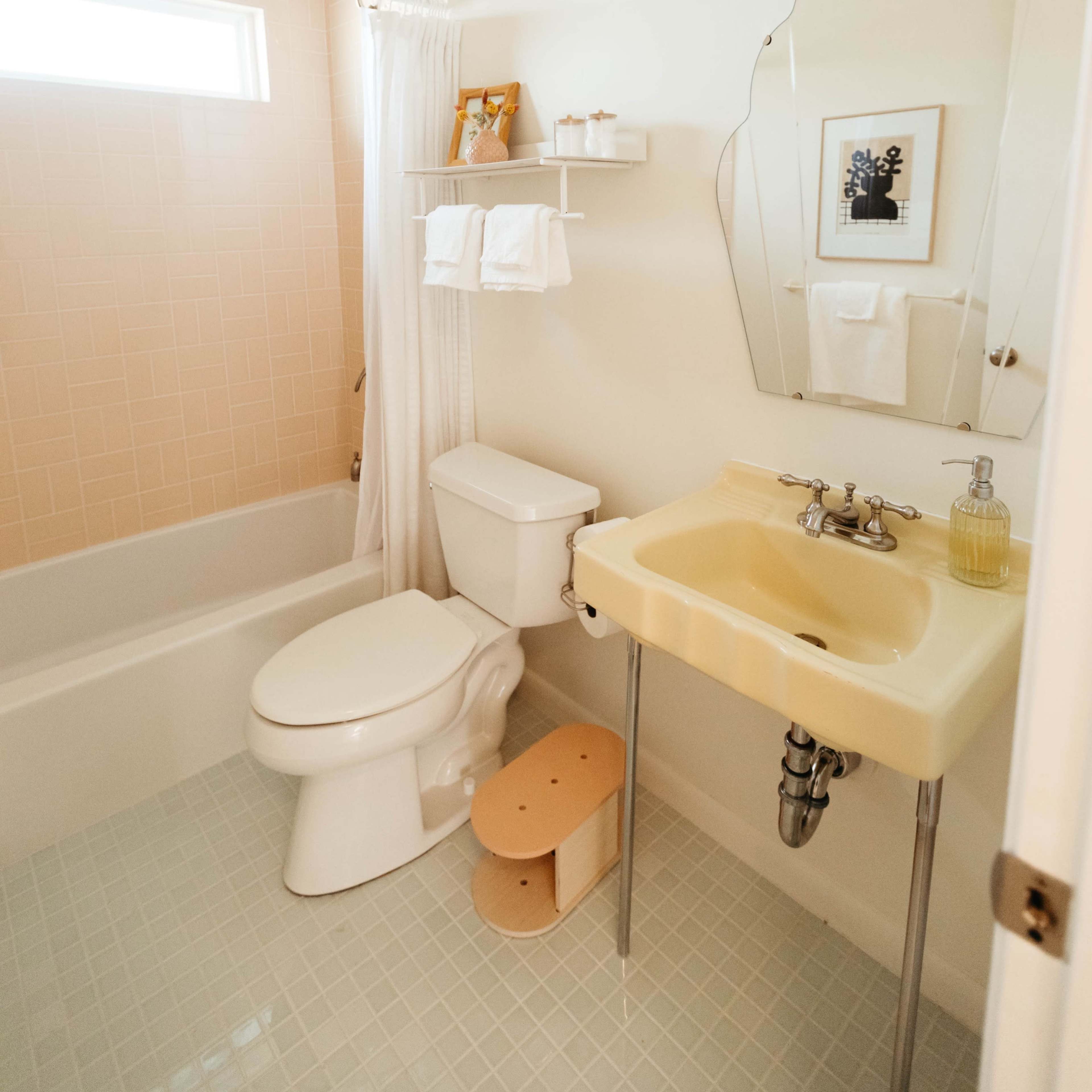 A small bathroom features a bathtub with a shower, a toilet, and a yellow sink with a mirror above it, all set against pale pink tiled walls.