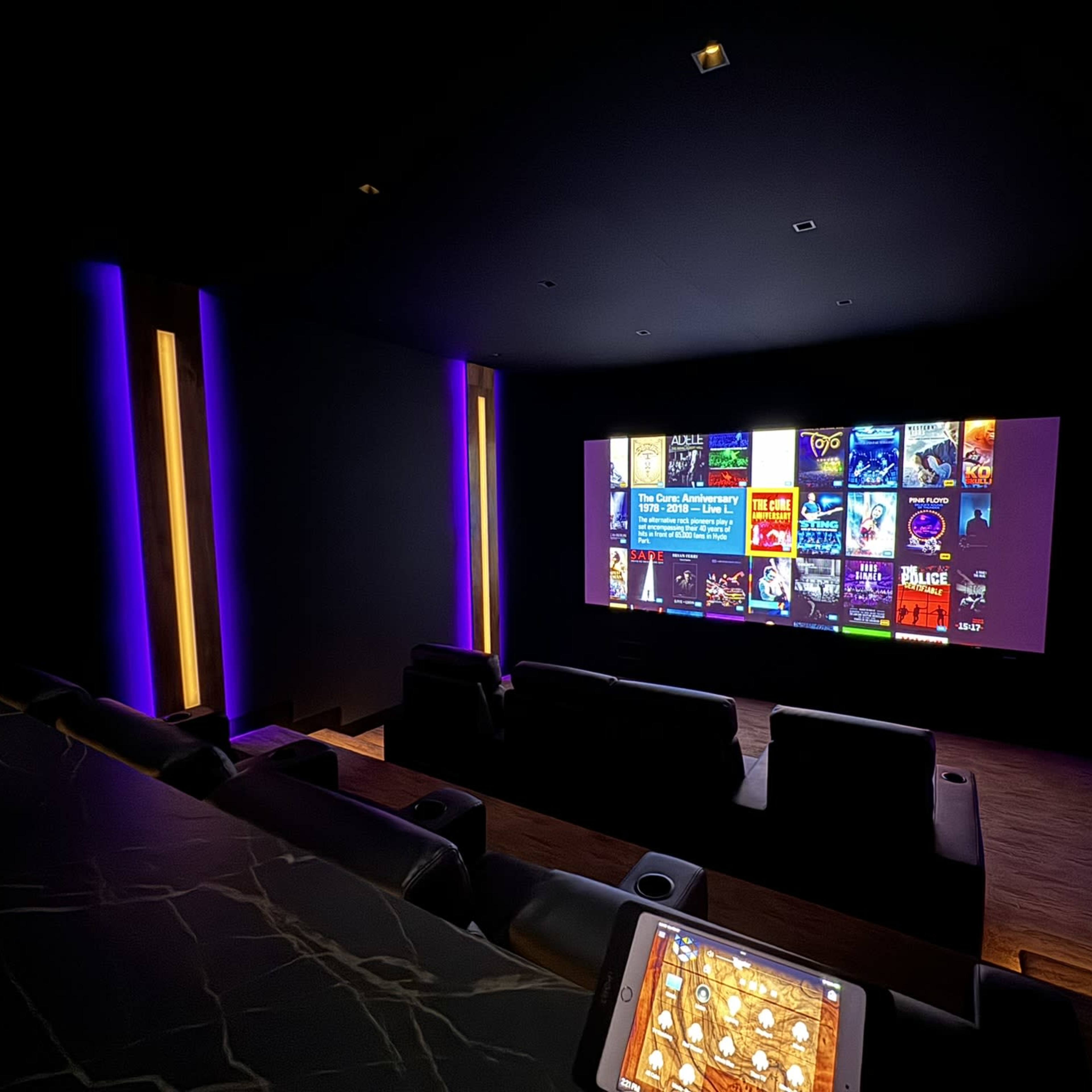 A dark home theater features a large screen displaying a movie selection interface, with leather seating and ambient lighting.