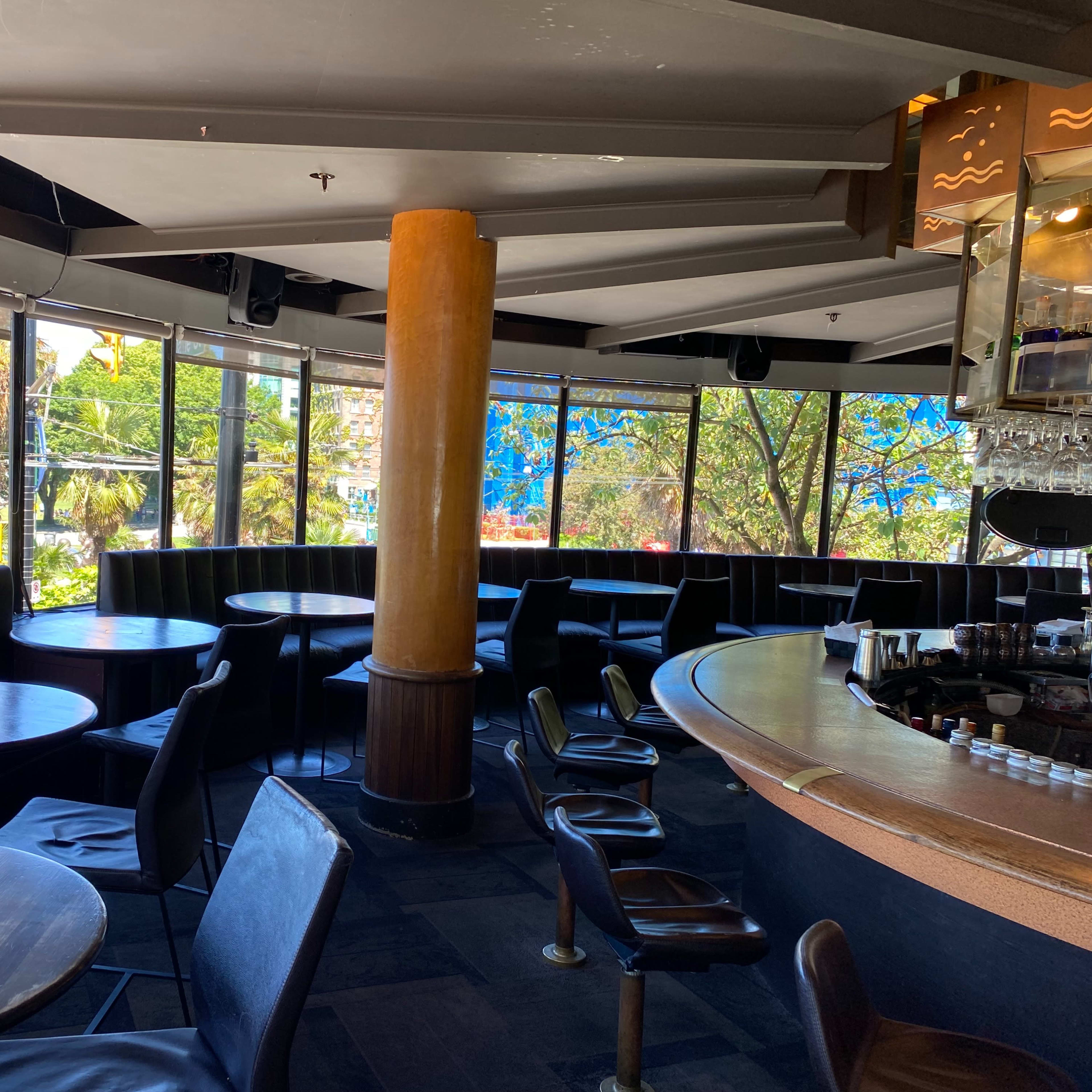 An empty bar area featuring round tables, black leather seating, and large windows overlooking a sunny outdoor scene.