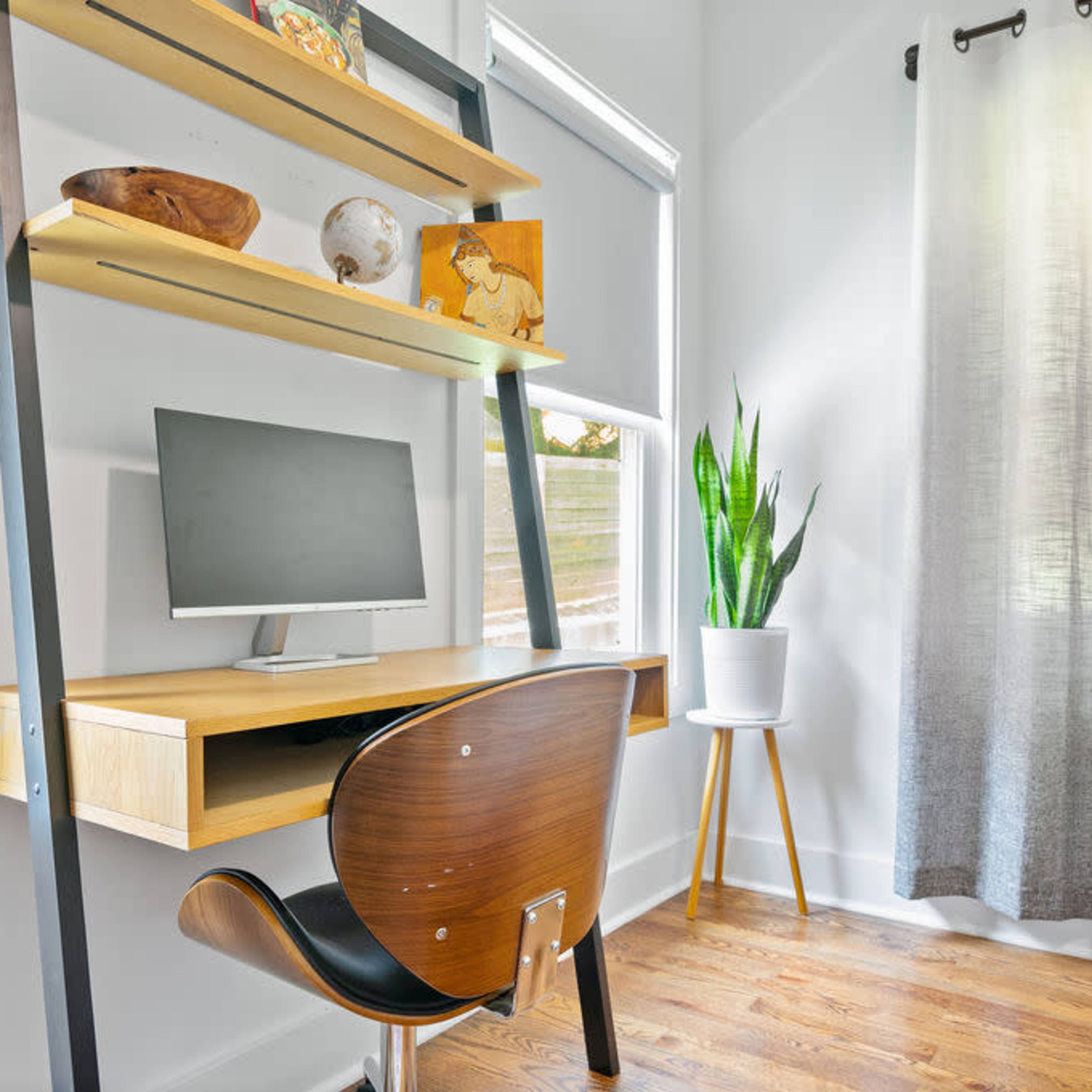 A minimalist home office features a wall-mounted desk with a computer monitor, an elegant wooden chair, shelves displaying decorative items, and a tall potted plant by the window.