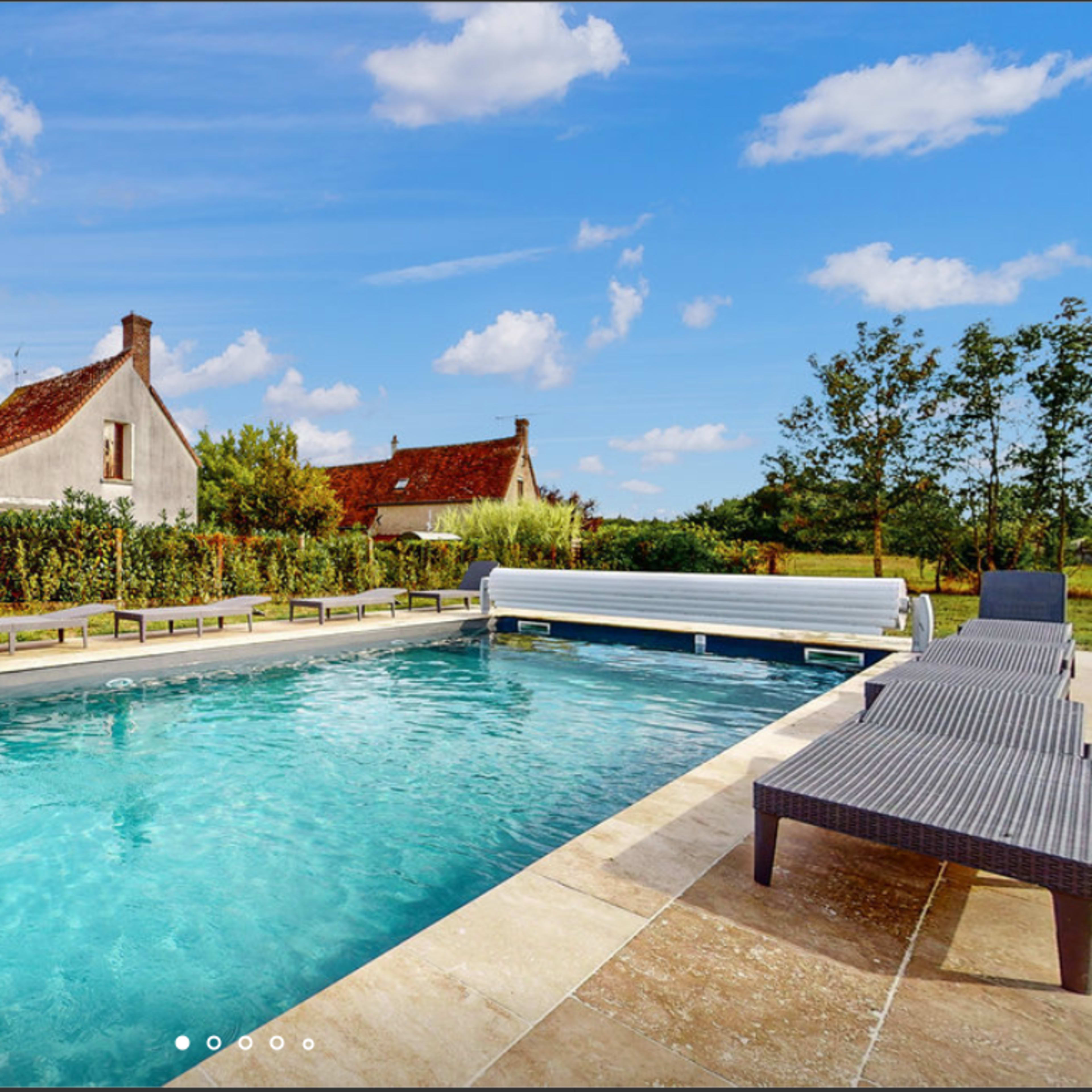 A clear swimming pool surrounded by a patio with loungers and a backdrop of rustic houses and greenery under a blue sky with scattered clouds.
