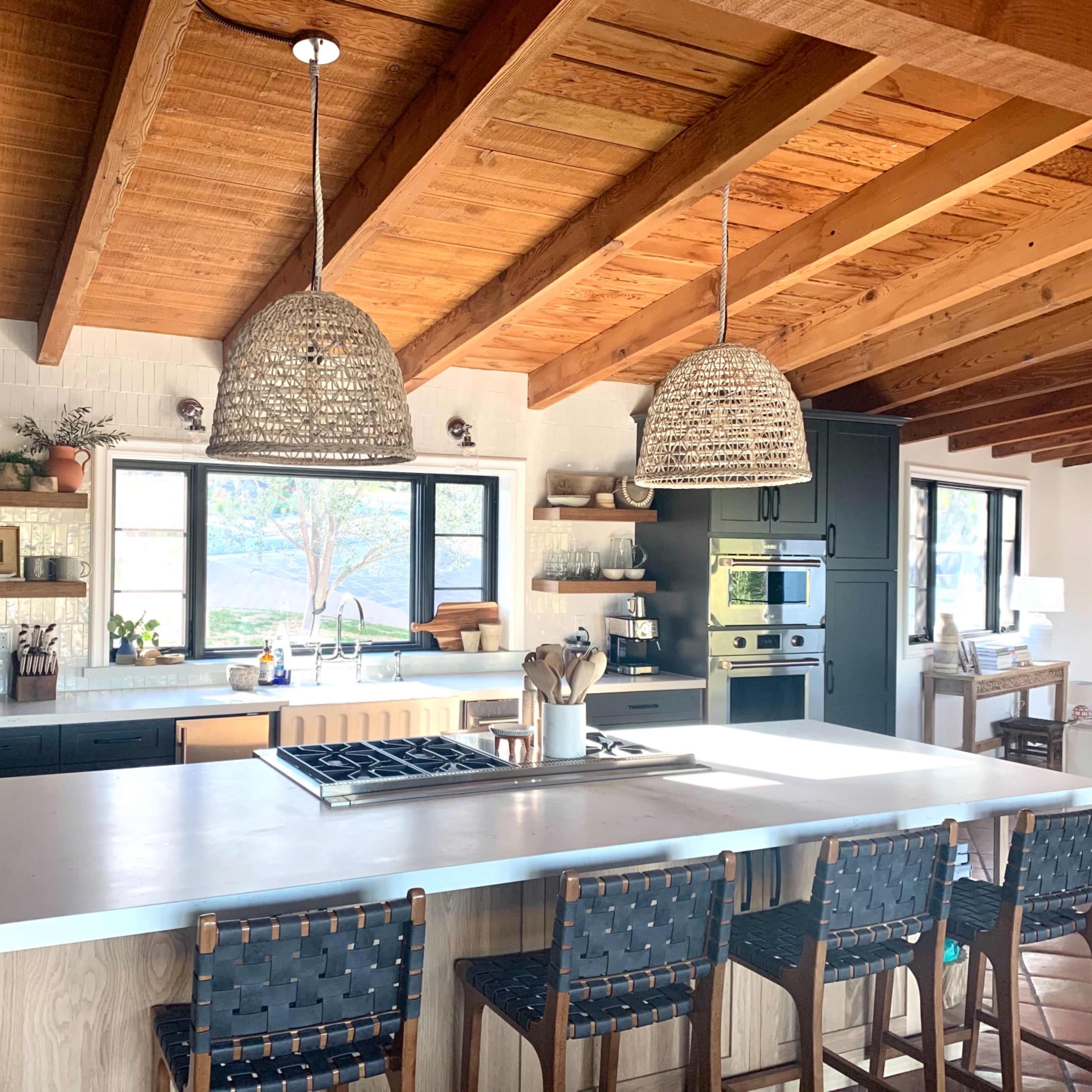 A modern kitchen features a large island with bar stools, woven pendant lights, and wooden ceiling beams.