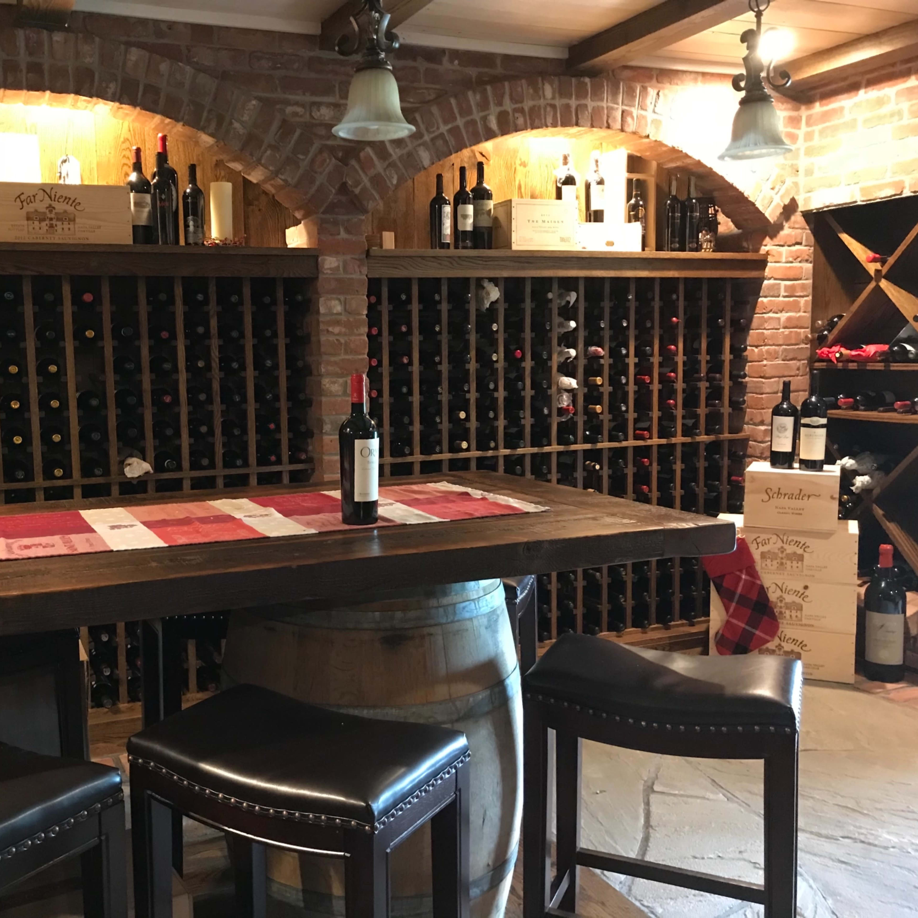 A rustic wine cellar featuring wooden wine racks filled with bottles, a bar table set with a wine bottle, and high-backed stools.