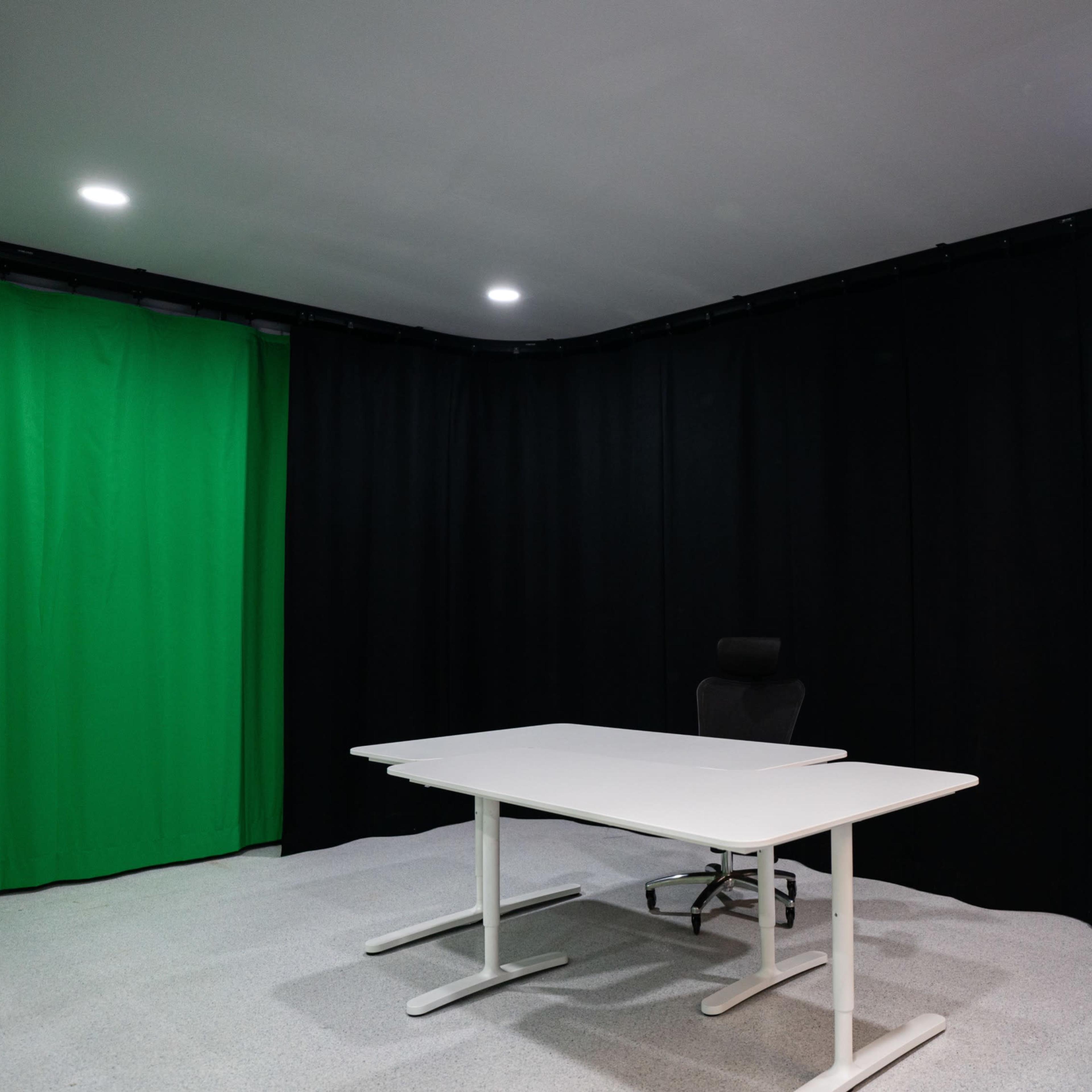 A video production studio with a white table, an office chair, and a green backdrop surrounded by black curtains.
