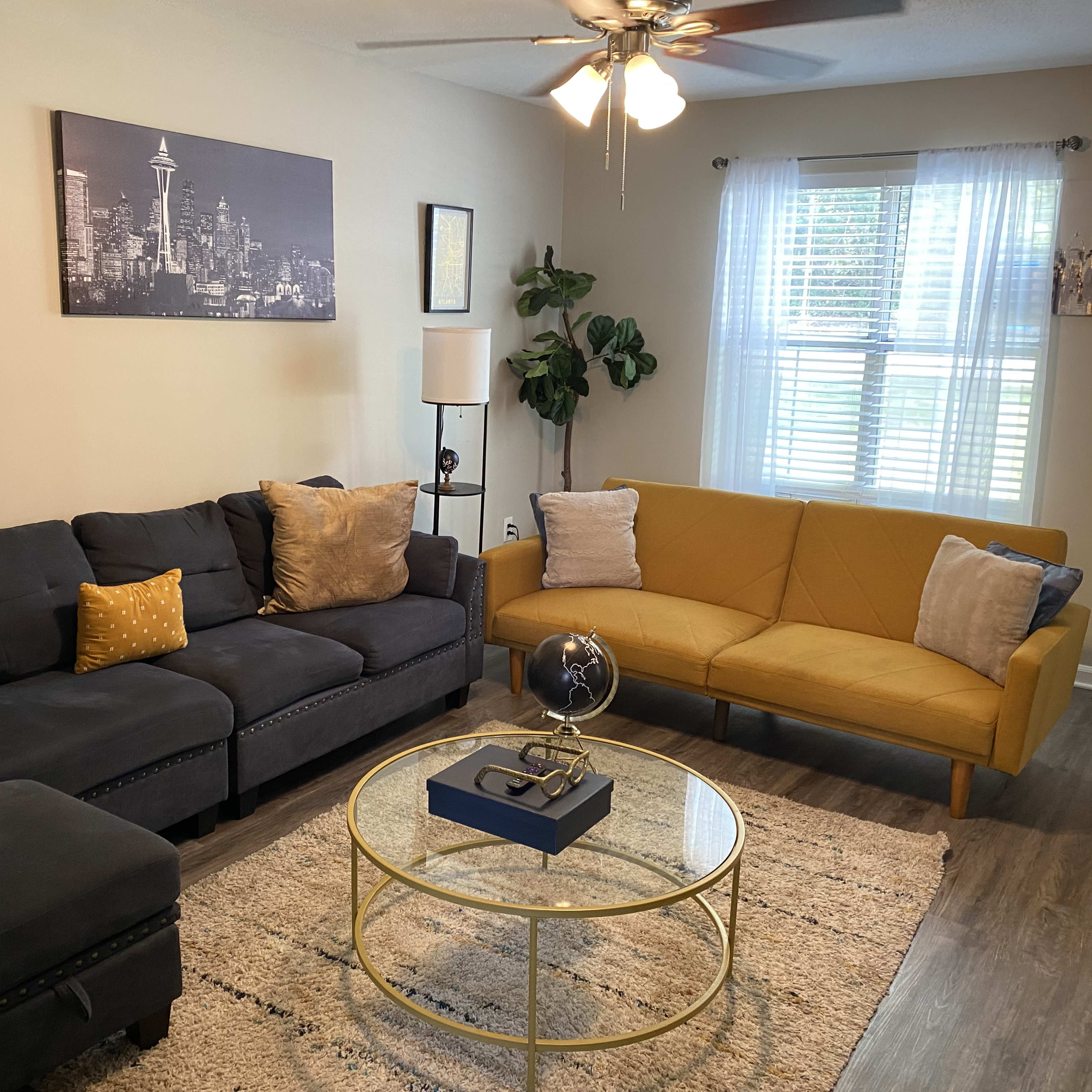 The living room features a dark sectional sofa, a bright yellow couch, and a round glass coffee table, with wall art and a plant enhancing the space.