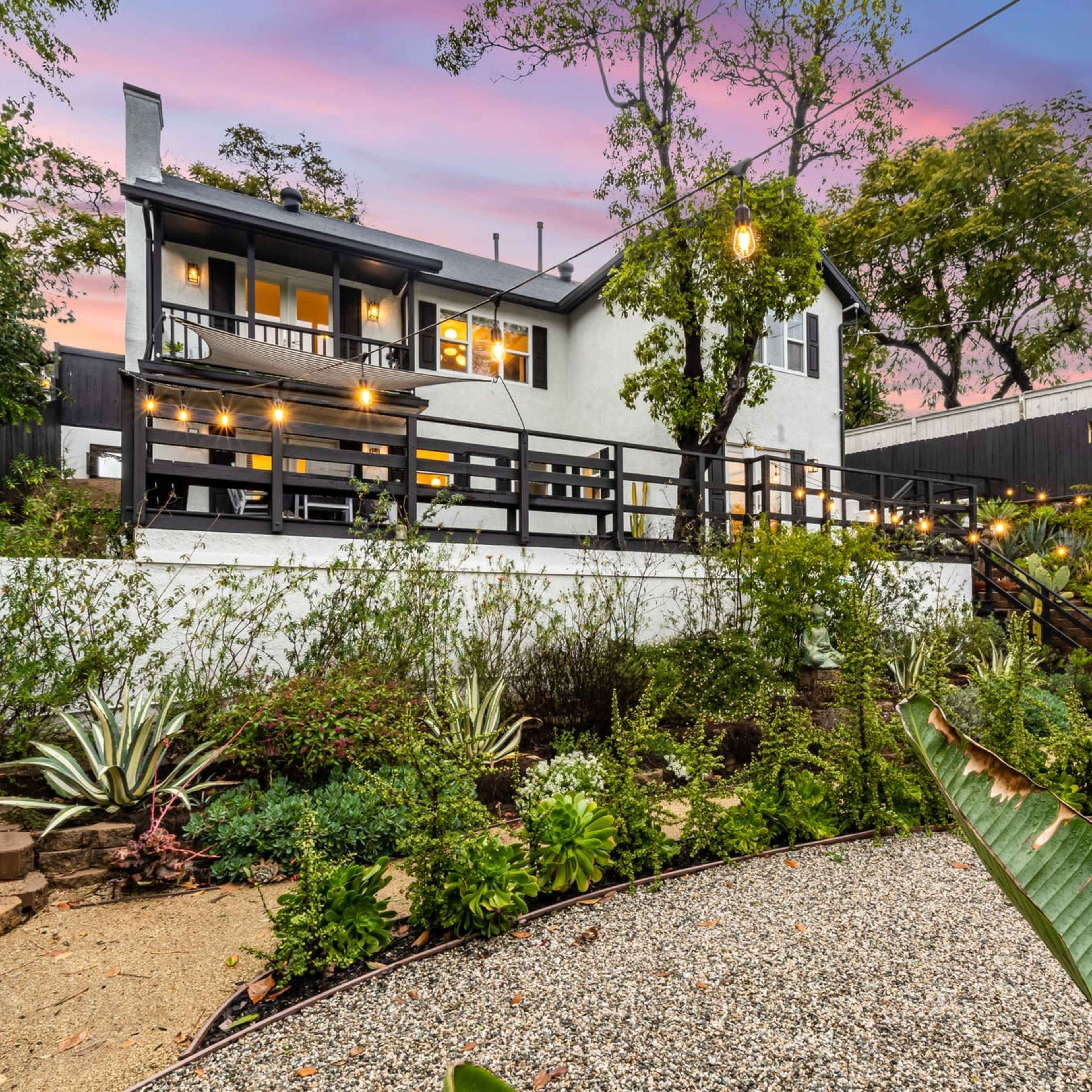 A white house with a black deck and string lights overlooks a landscaped garden with various plants and a gravel path.