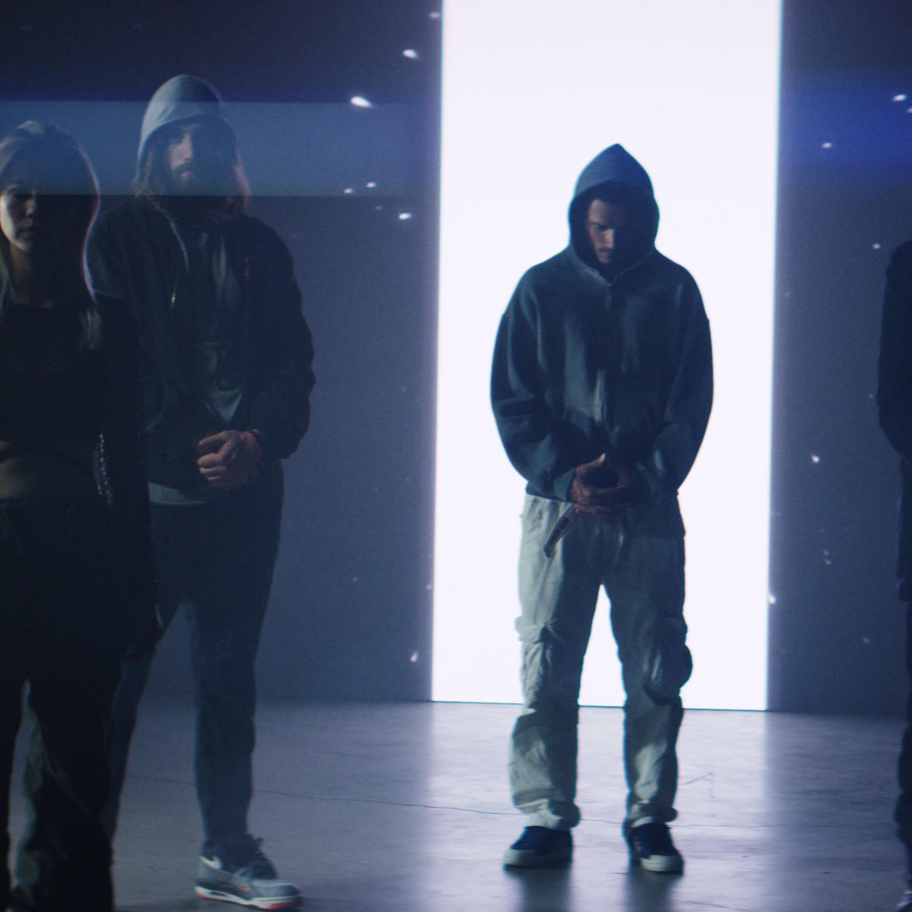 A group of five individuals in hooded attire stands in a dimly lit space with a bright vertical light behind them.