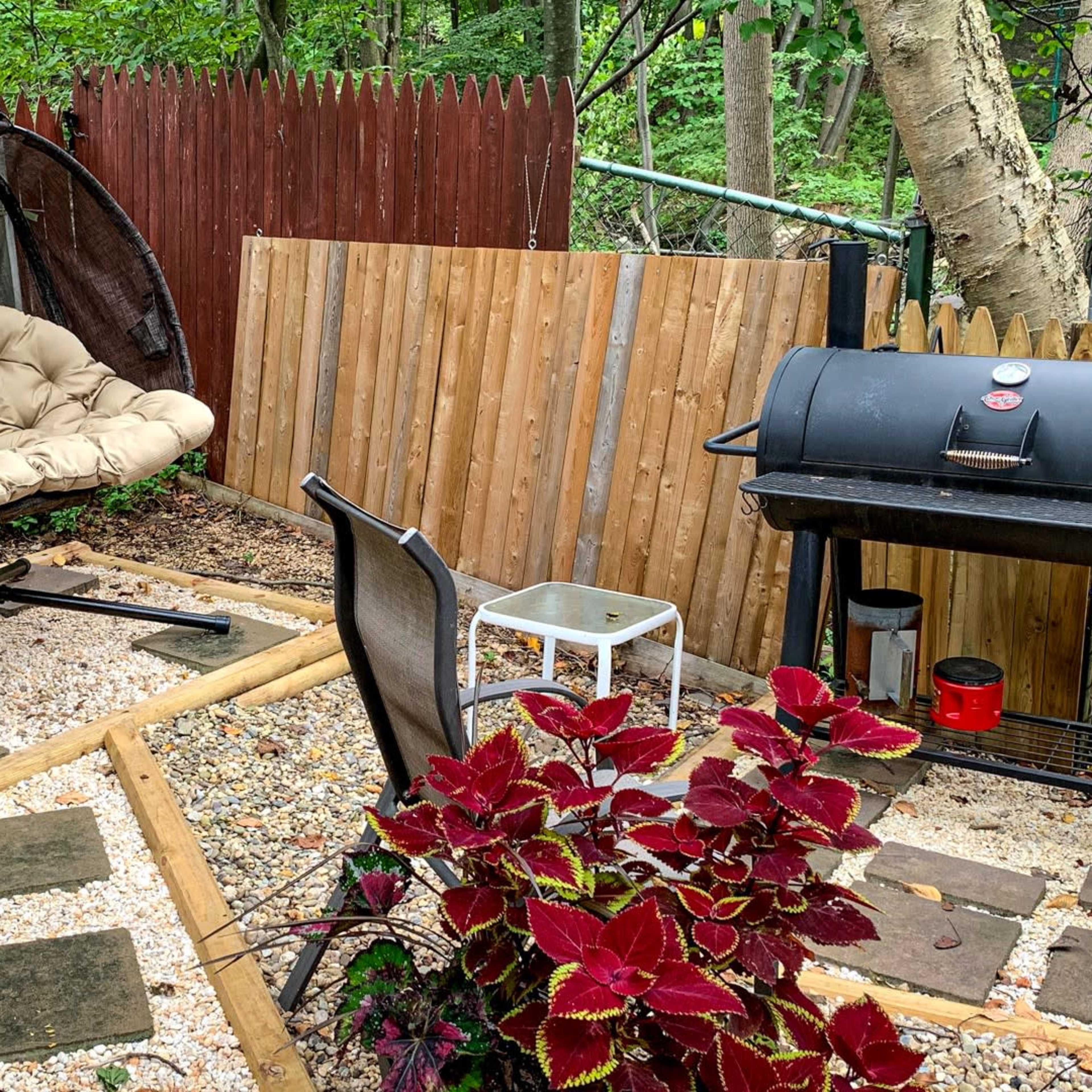 A small outdoor area featuring a hanging chair, a grill, gravel pathways, and a potted plant.