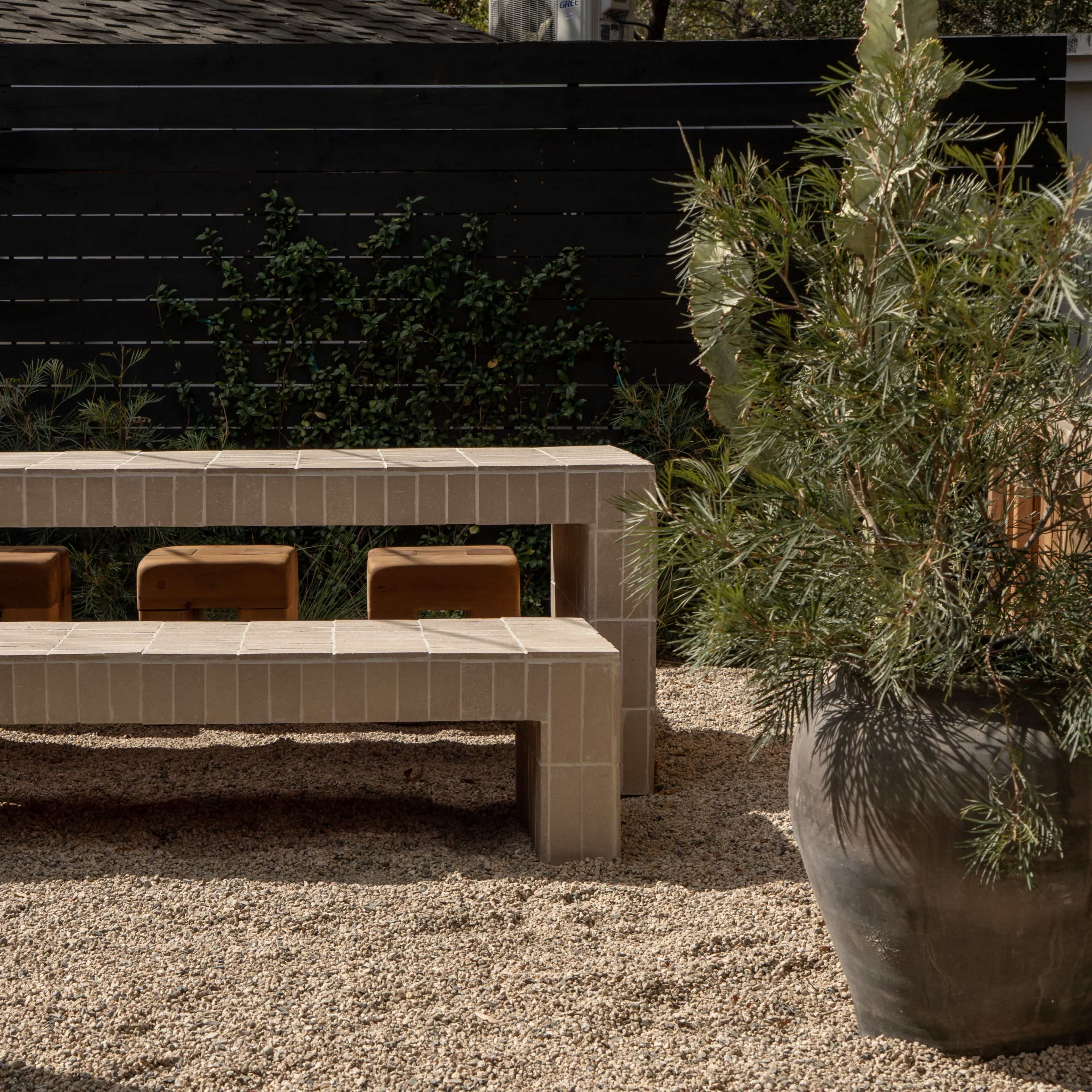 A stone bench and table set in a gravel garden surrounded by potted plants and a dark wooden fence.