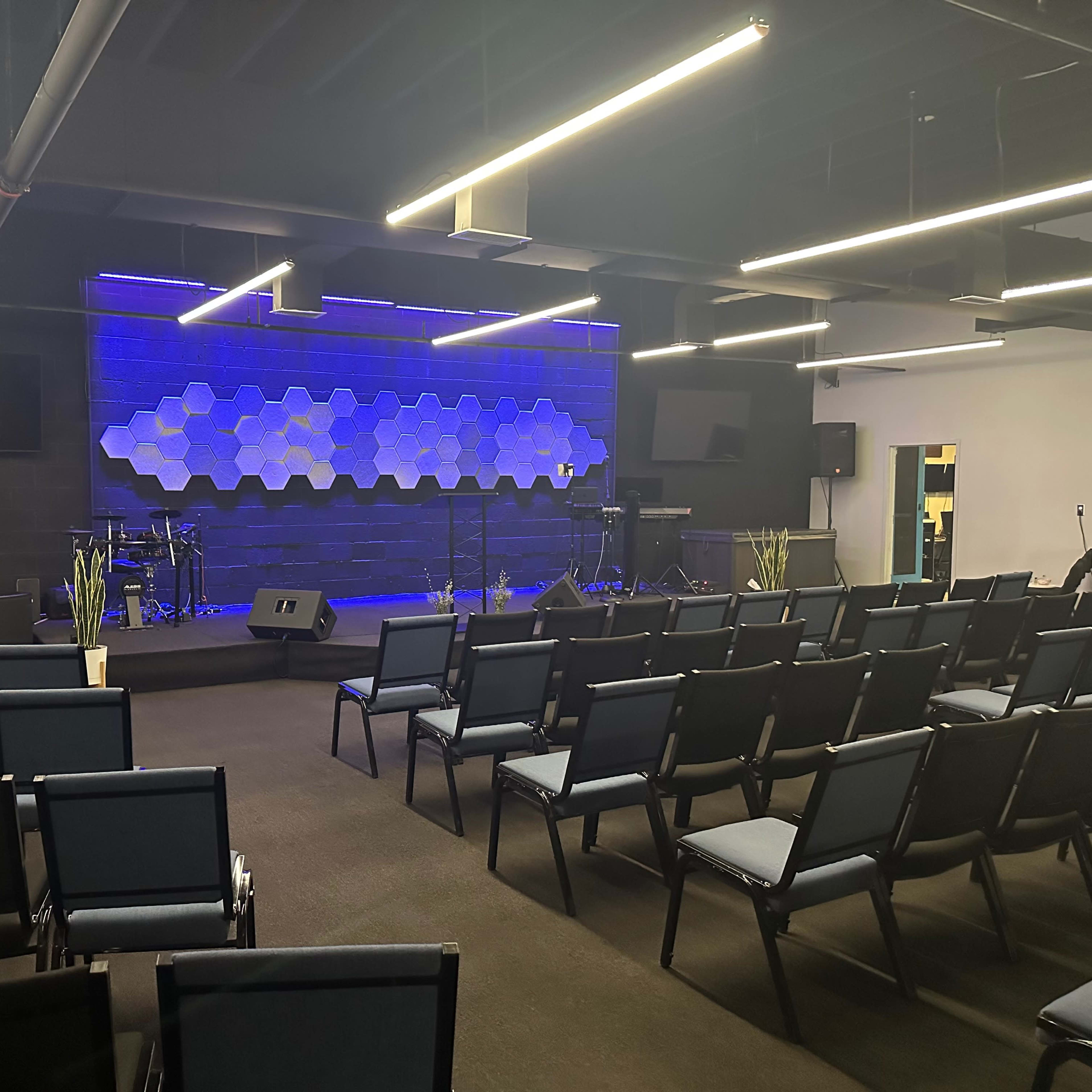 A modern interior of a performance space with rows of chairs facing a stage featuring a hexagonal backdrop, musical instruments, and lighting.