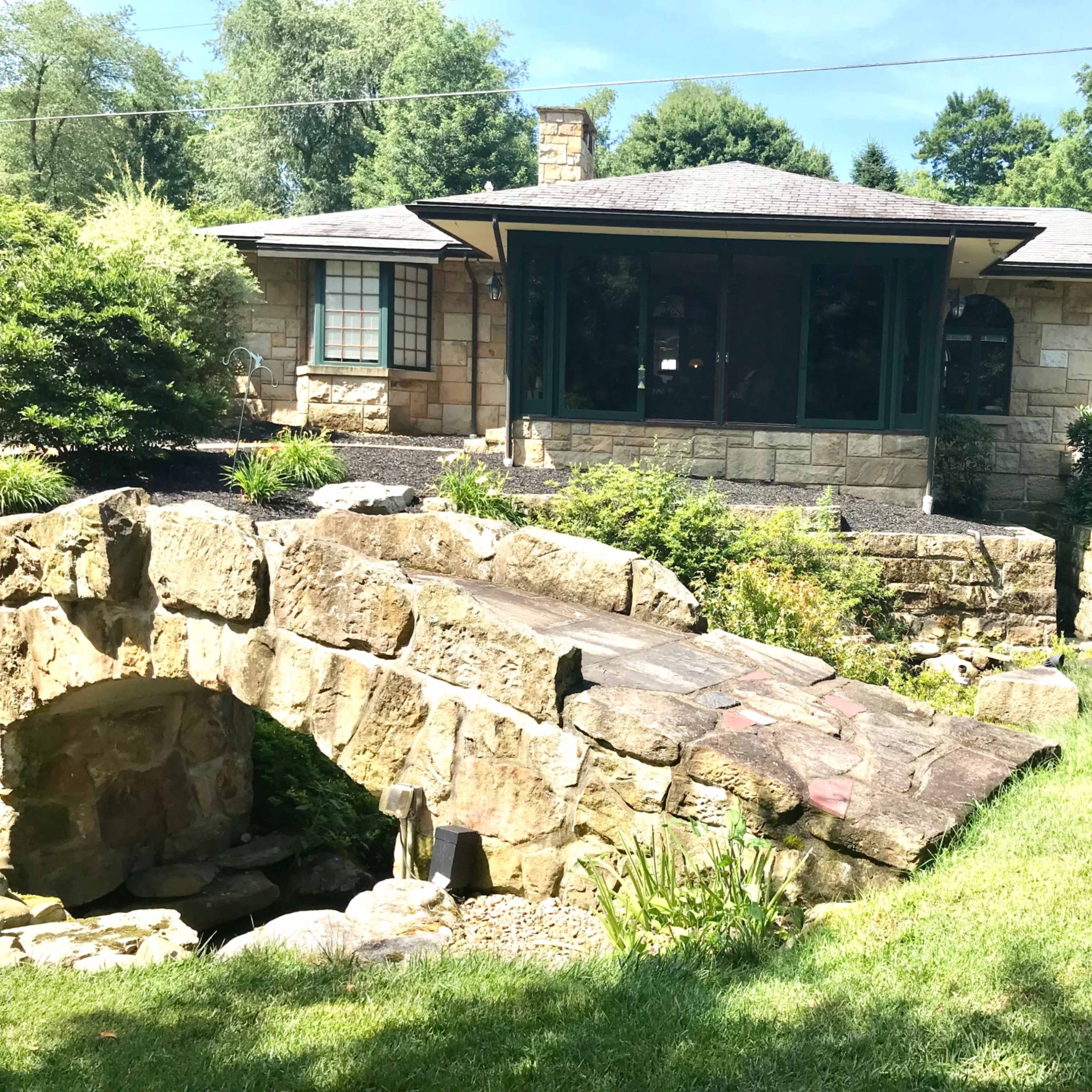A stone house with a large porch and a small stone bridge over a landscaped garden.