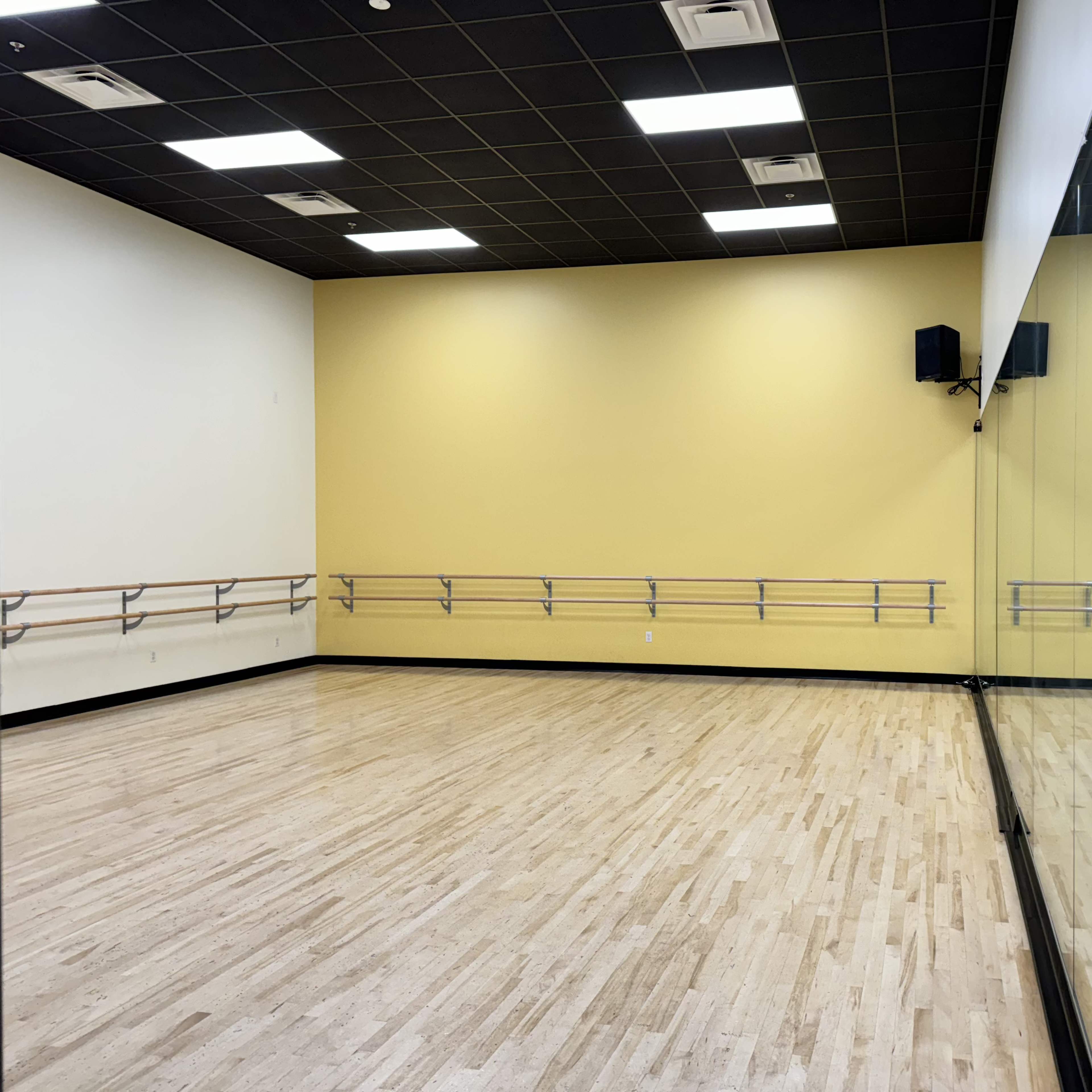 A spacious dance studio with wooden flooring, yellow walls, and a mirrored wall along one side.