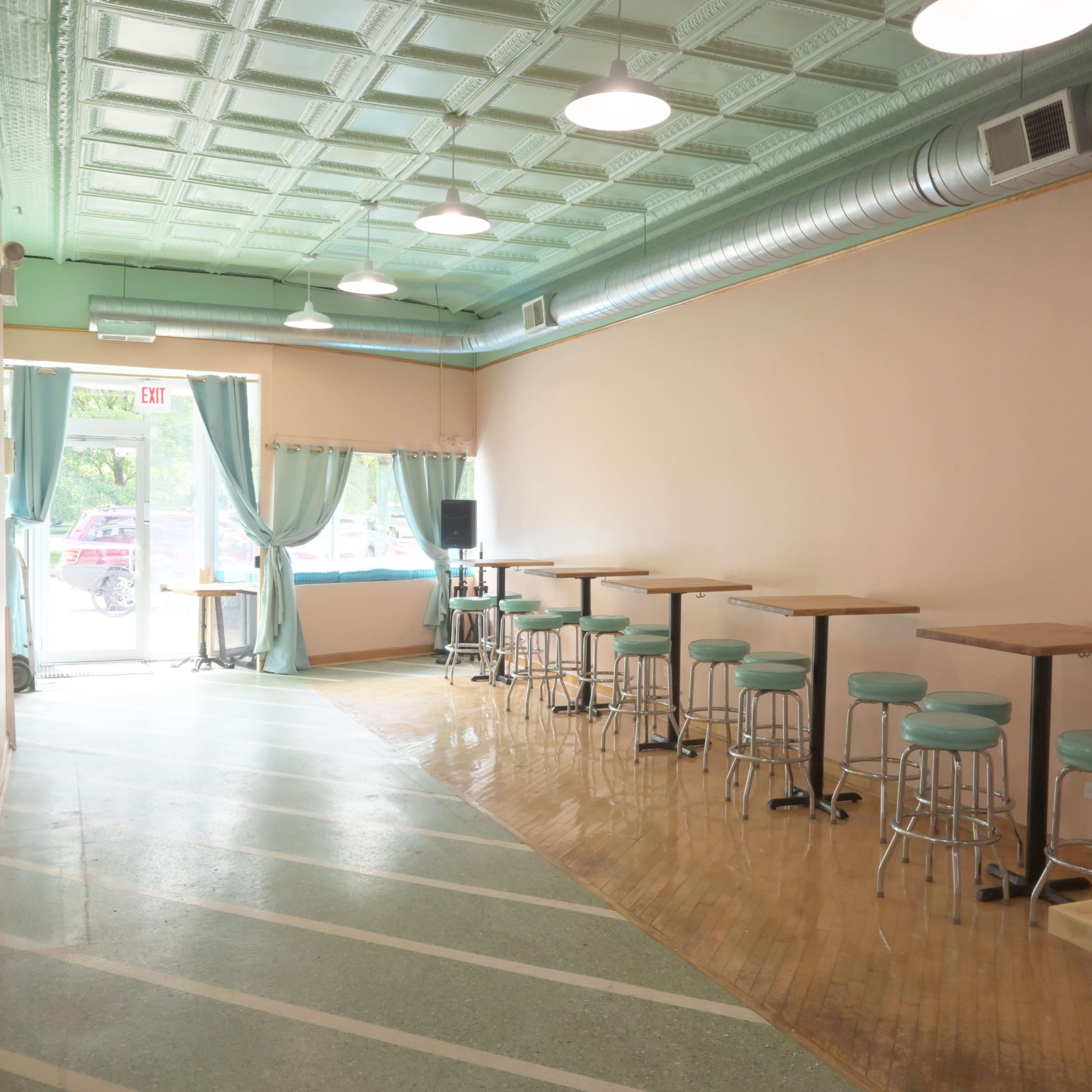 A narrow, vacant café interior with a light pink wall, metal stools along the side, and large windows allowing natural light to enter.