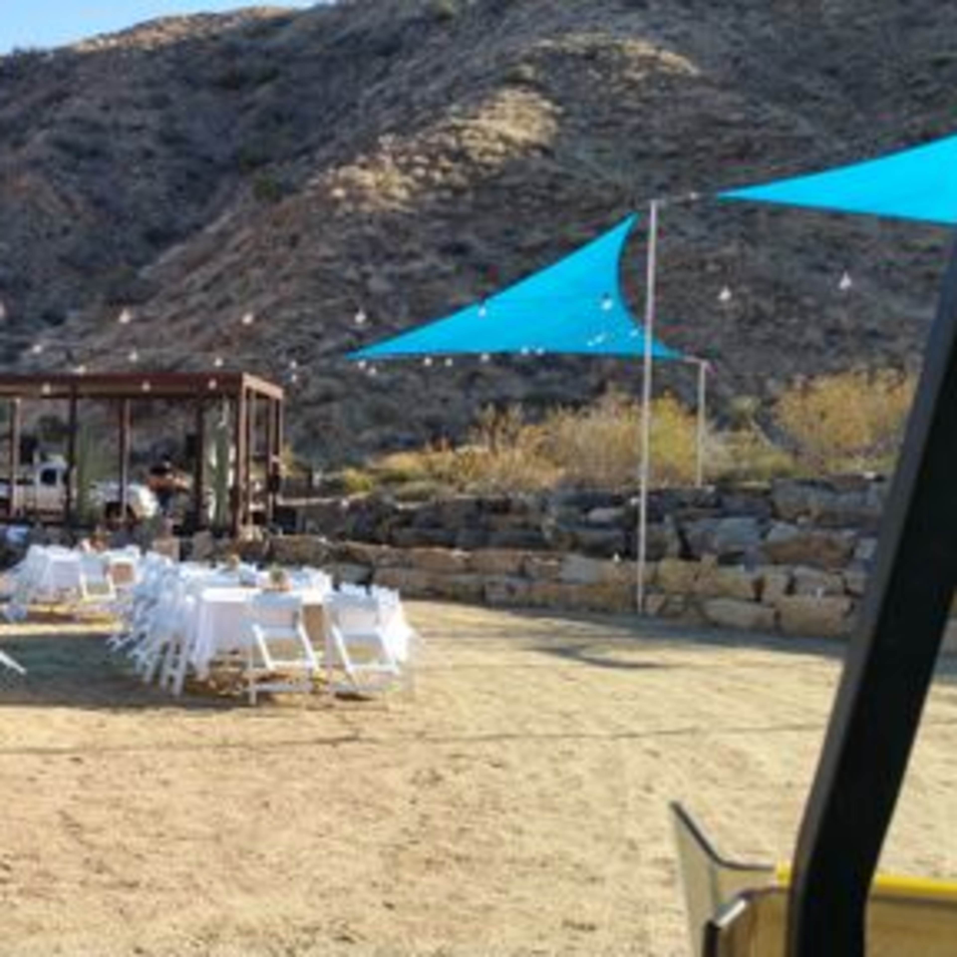 An outdoor event space with white tables and chairs arranged under blue canopies, near a food truck and a wooden structure in a desert landscape.