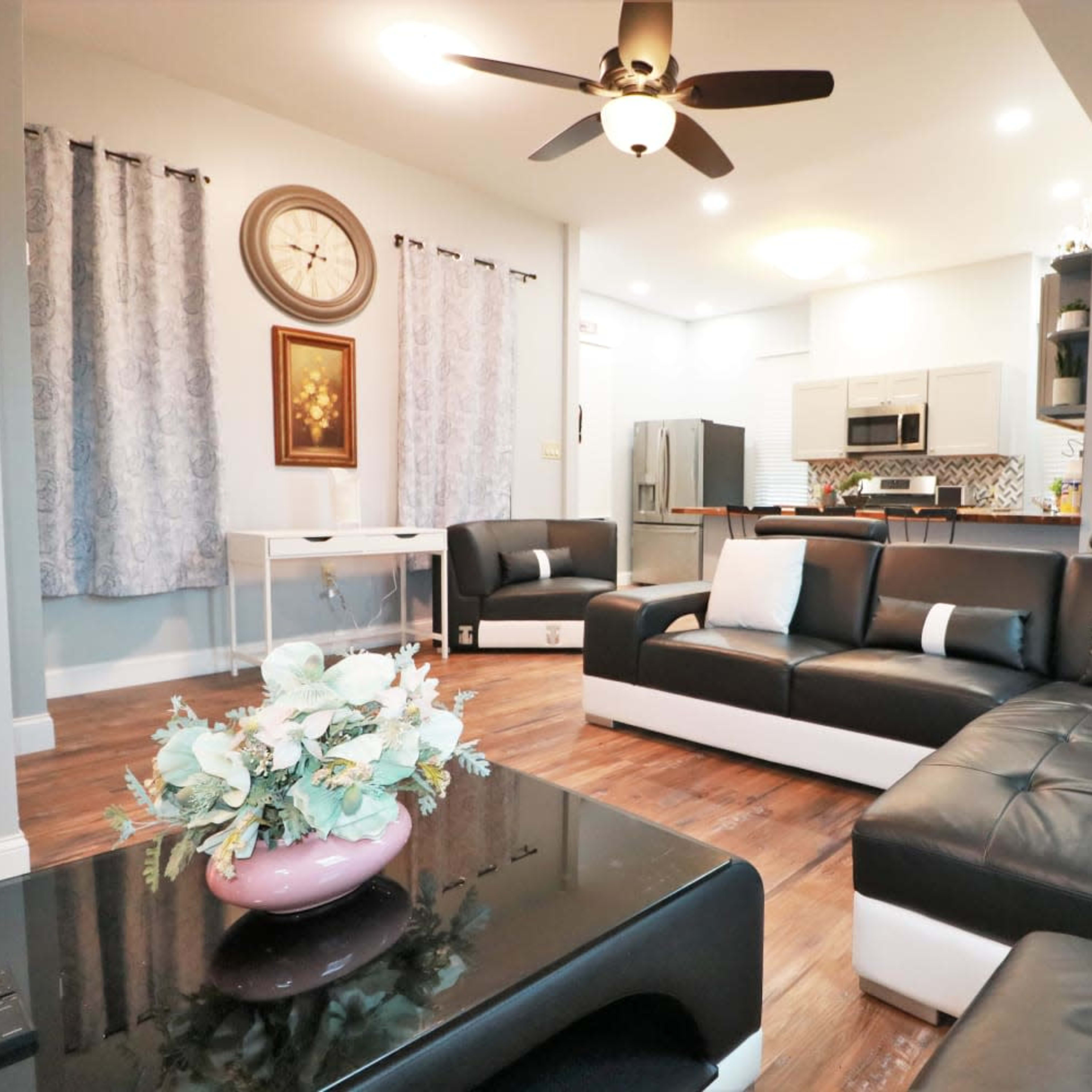A modern living room with black leather furniture, a coffee table, decorative curtains, and a kitchen area in the background.