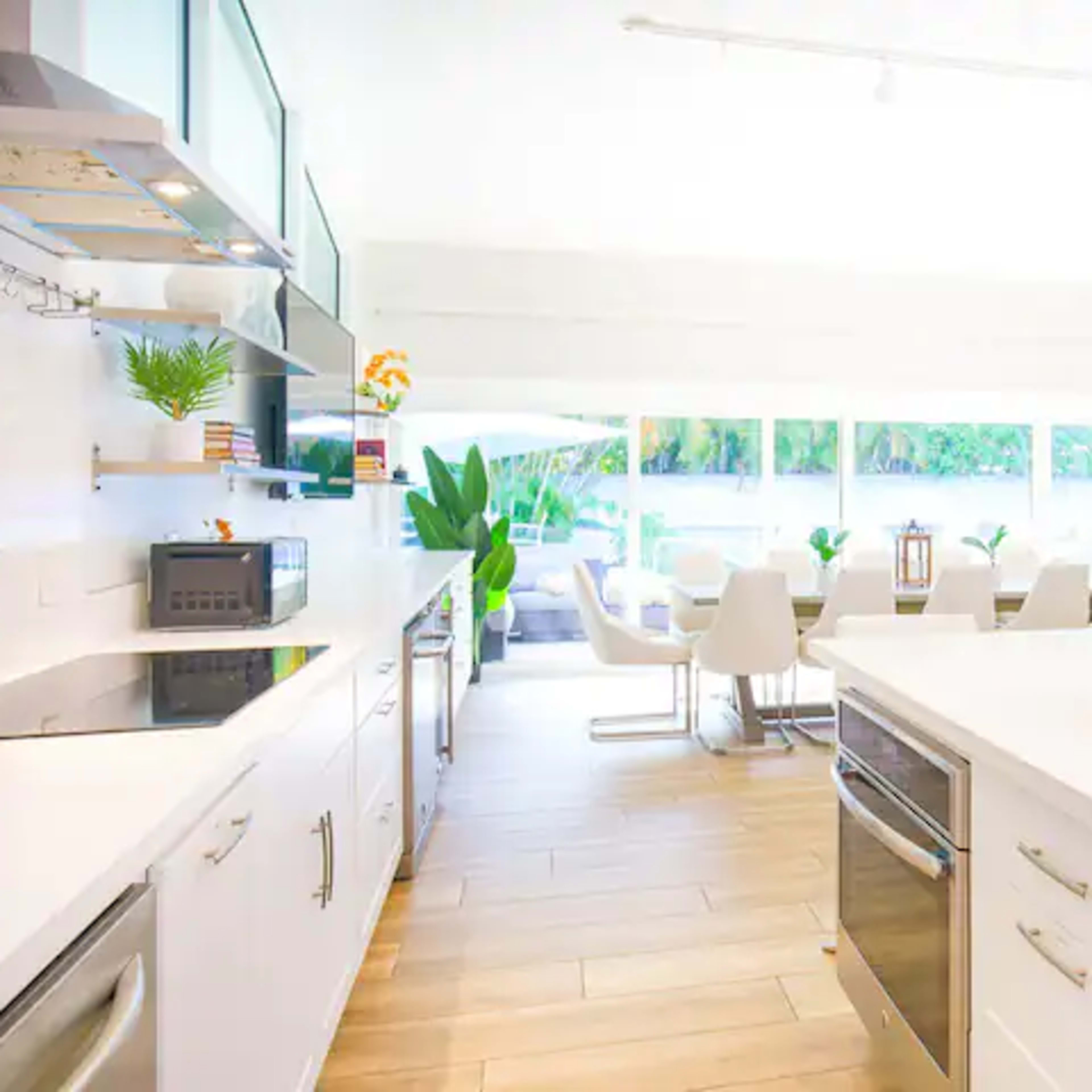 A modern kitchen with white cabinetry, stainless steel appliances, and a dining area featuring white chairs and large windows that provide ample natural light.
