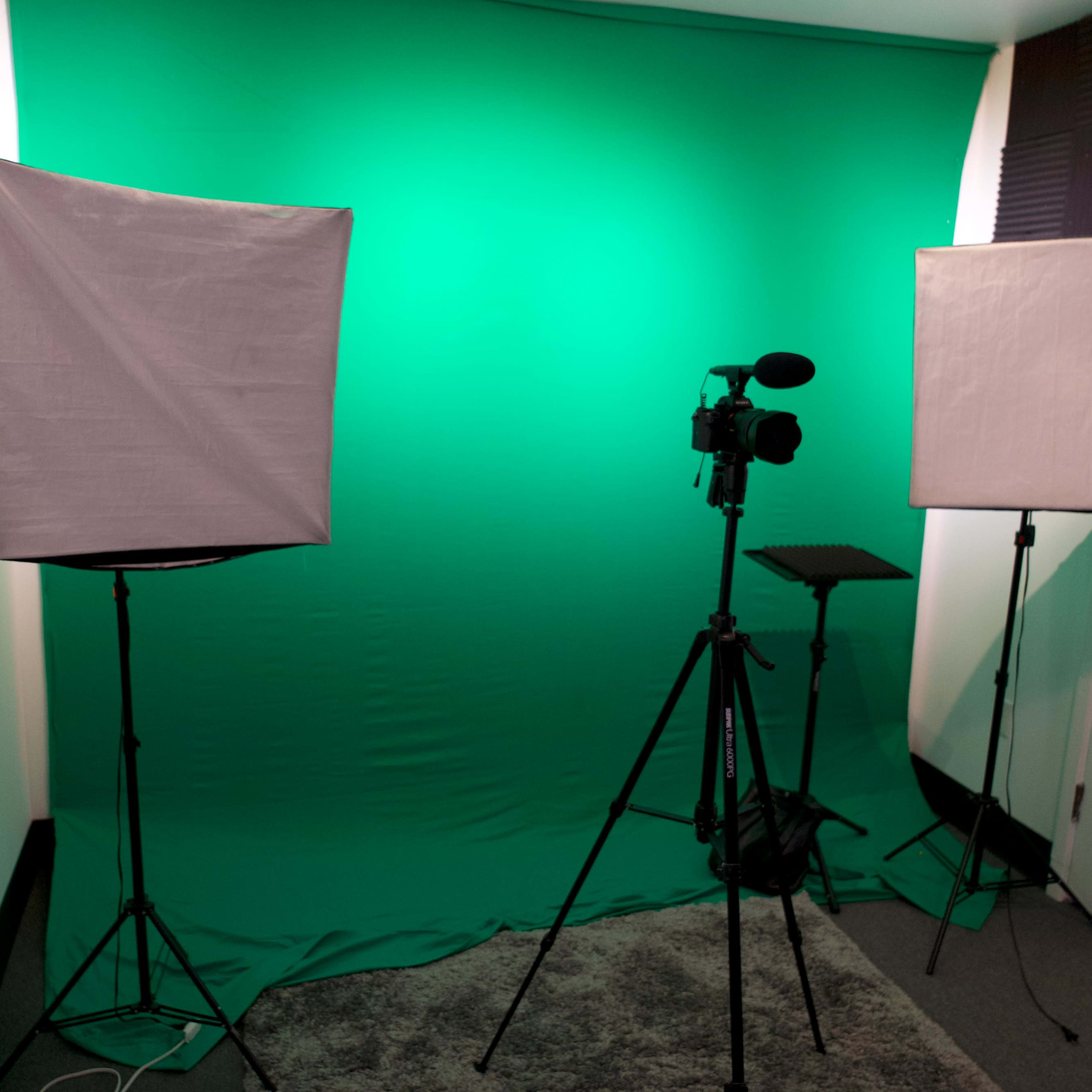 A room equipped with a green screen backdrop, two softbox lights, and a camera on a tripod.