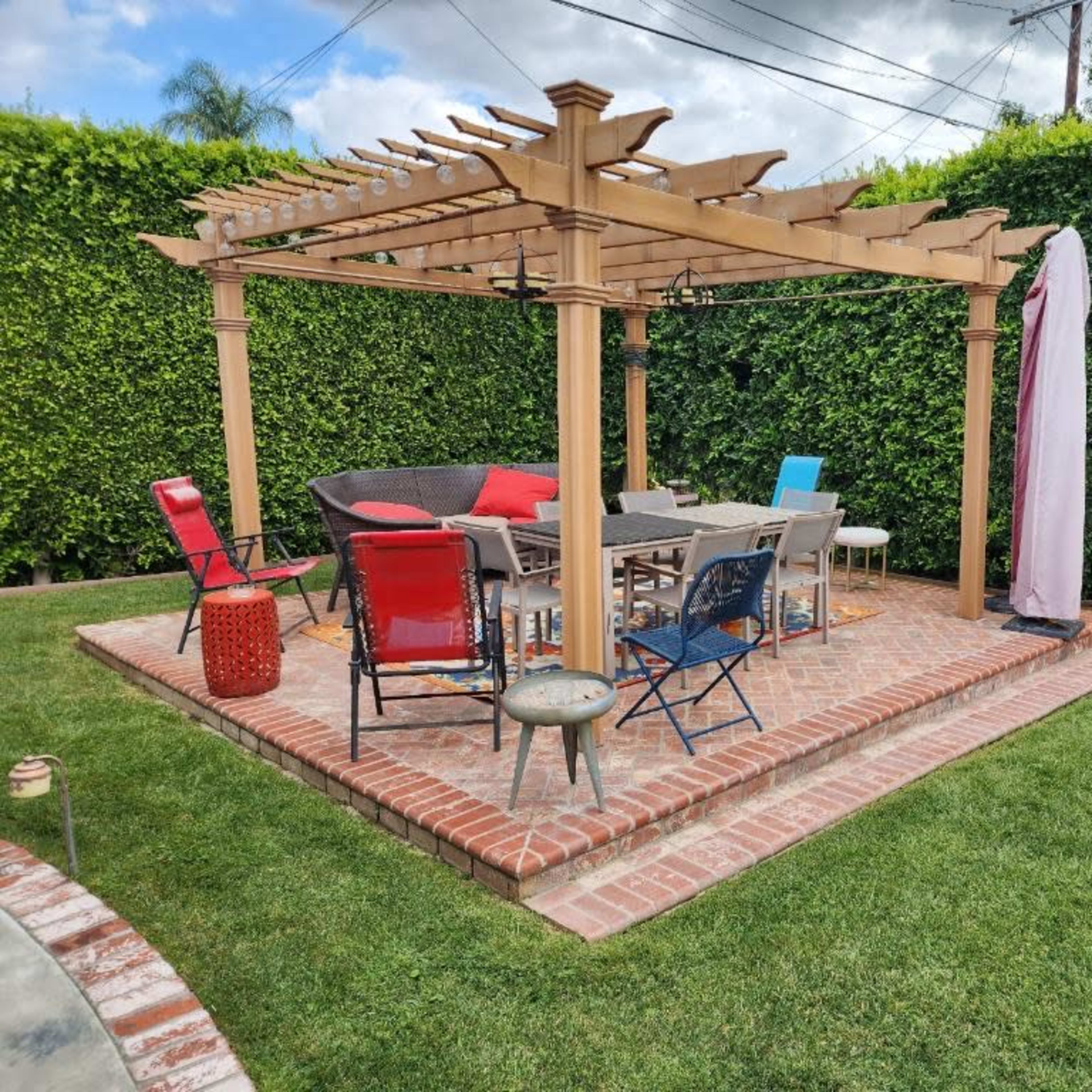A outdoor patio features a wooden pergola with seating arrangements, surrounded by green hedges and brick pavement.